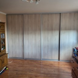 Krystian Szkotak - Duża szafa przesuwna z jasnego drewna z czterema drzwiami, zabudowana od ściany do ściany, widoczna na tle parkietu i fragmentów mebli.