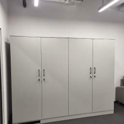 Krystian Szkotak - Biała, minimalistyczna szafa z czterema drzwiami i czarnymi uchwytami, z kluczami w zamkach, na tle szarej wykładziny w nowoczesnym biurze z widocznymi elementami wentylacji pod sufitem.