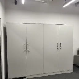 Biała, minimalistyczna szafa z czterema drzwiami i czarnymi uchwytami, z kluczami w zamkach, na tle szarej wykładziny w nowoczesnym biurze z widocznymi elementami wentylacji pod sufitem.