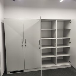 Krystian Szkotak - Biała szafa biurowa z dwiema parami drzwi zamykanymi na klucz i otwartą częścią regałową z półkami, widoczna instalacja wentylacyjna pod sufitem.