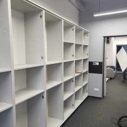 Krystian Szkotak - Biała, modułowa konstrukcja regałowa z otwartymi półkami, widoczne zawiasy, w tle osoba przenosząca białą płytę, szara wykładzina podłogowa i fragment odkurzacza.