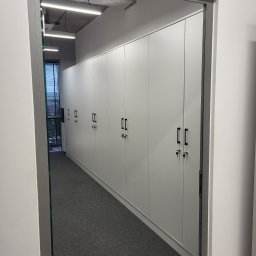 Krystian Szkotak - Długi rząd białych szaf z czarnymi uchwytami i zamkami, widoczny zza framugi drzwiowej w biurze z szarą wykładziną.