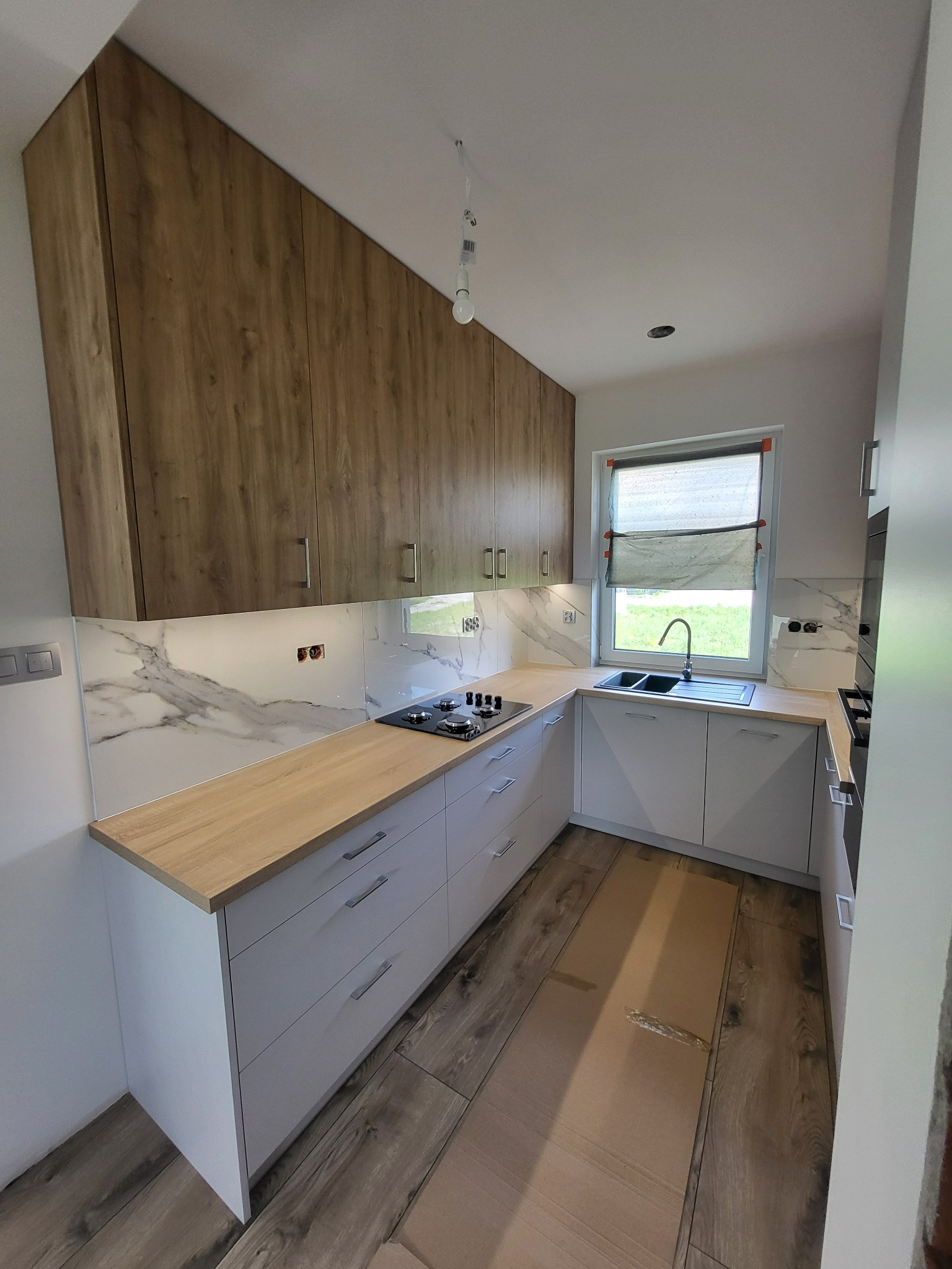 Nowoczesna kuchnia z białymi szafkami dolnymi i drewnianymi górnymi, marmurowy backsplash, drewniany blat i podłoga, częściowo zakryte okno.