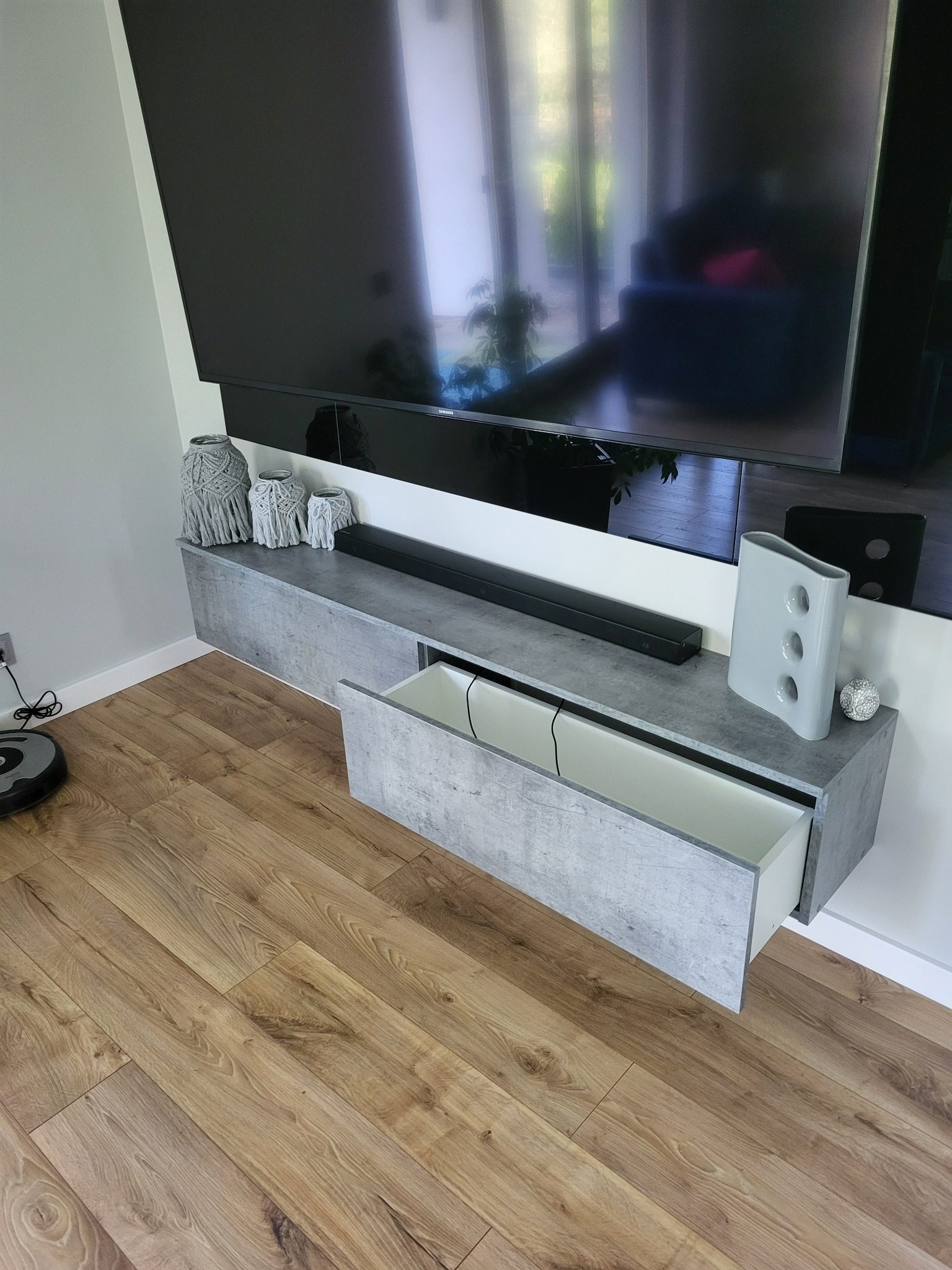 Szara szafka RTV z otwartą szufladą, dekoracjami makrama i wazonem, zawieszona pod telewizorem, z robotem sprzątającym na parkiecie.