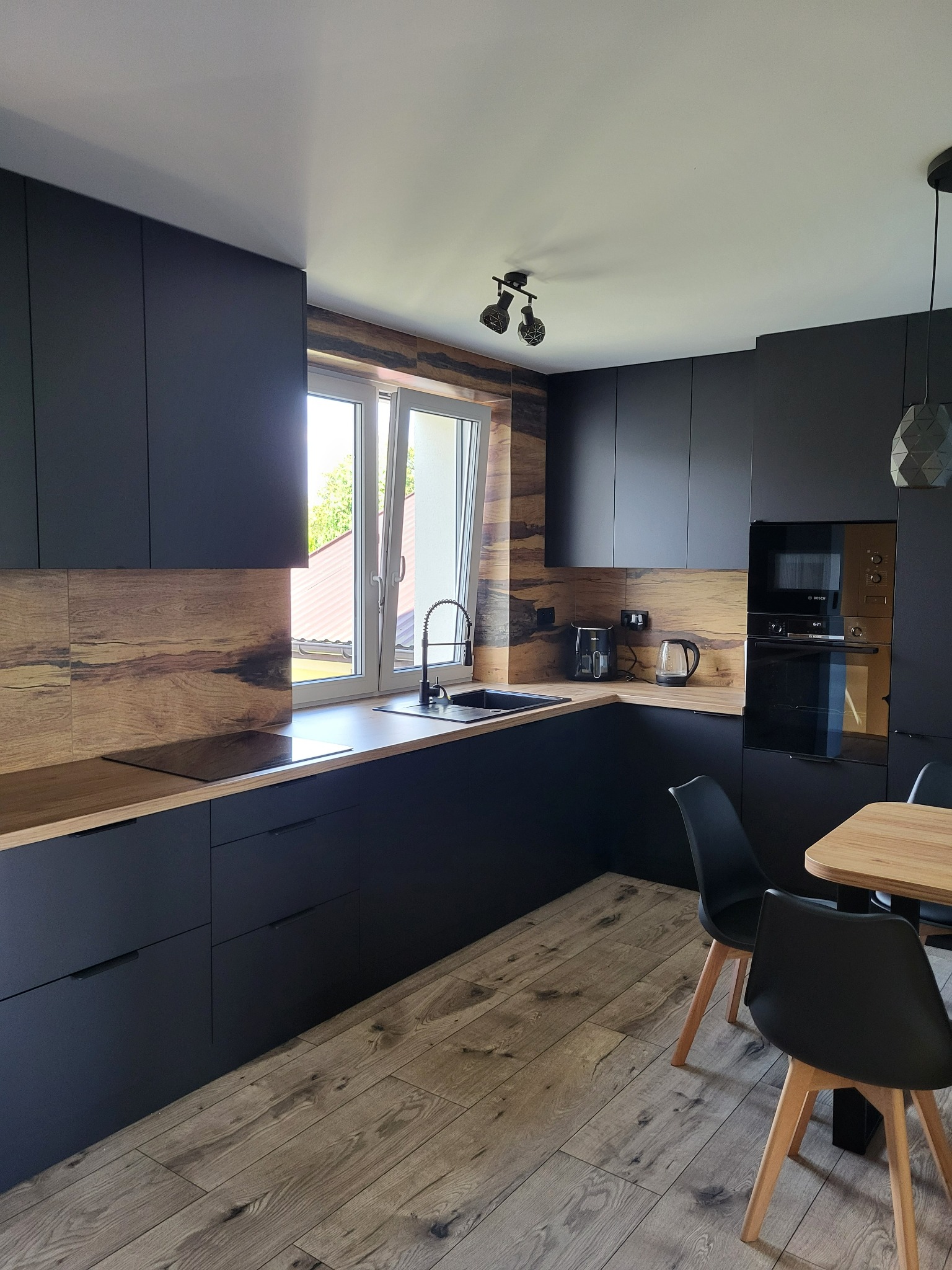 Nowoczesna kuchnia z czarnymi matowymi frontami, drewnianym blatem i panelami ściennymi o wzorze drewna, widok na okno i stół z krzesłami.