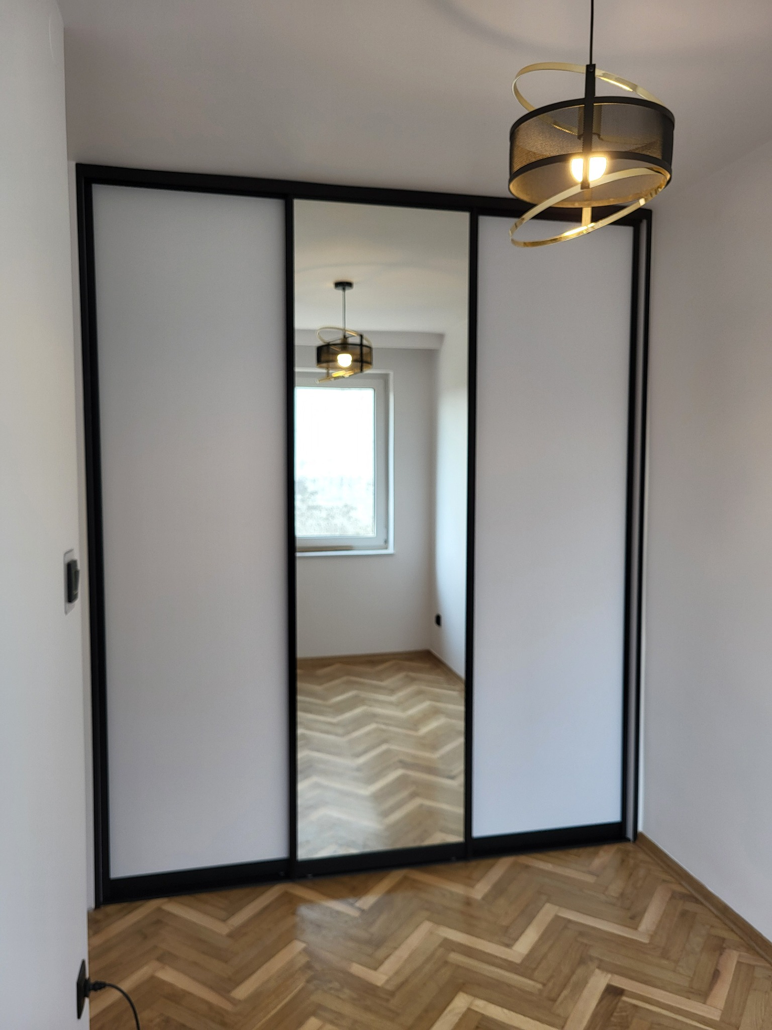 Trzydrzwiowa szafa z przesuwnymi drzwiami: dwie białe matowe płyty po bokach i lustro pośrodku w czarnej ramie, parkiet w jodełkę na podłodze, lampa sufitowa z czarnym abażurem i złotymi elementami.