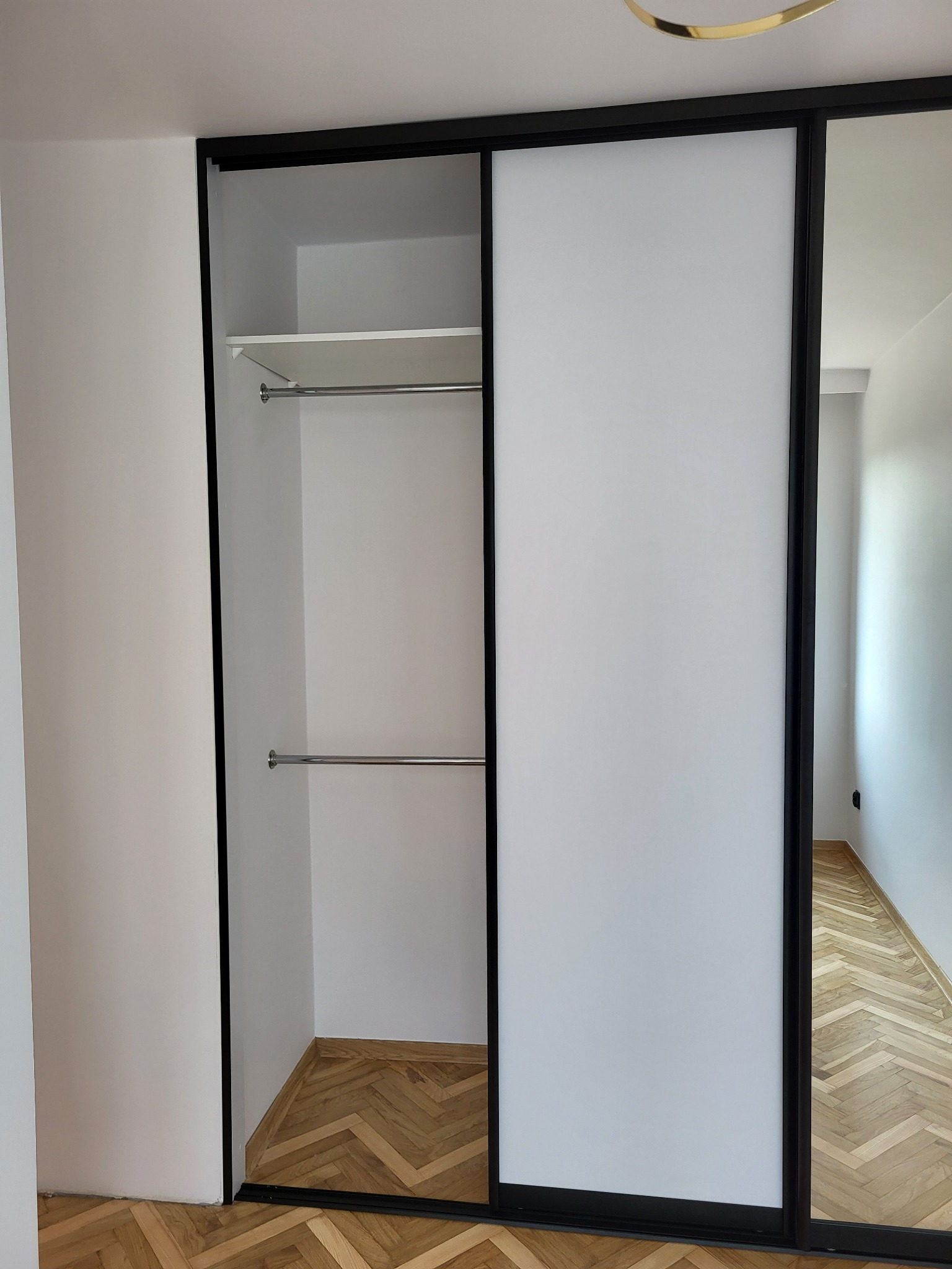 Biała szafa przesuwna z czarnymi ramami, z widocznym wnętrzem z półką i dwoma drążkami na ubrania, na podłodze parkiet ułożony w jodełkę.