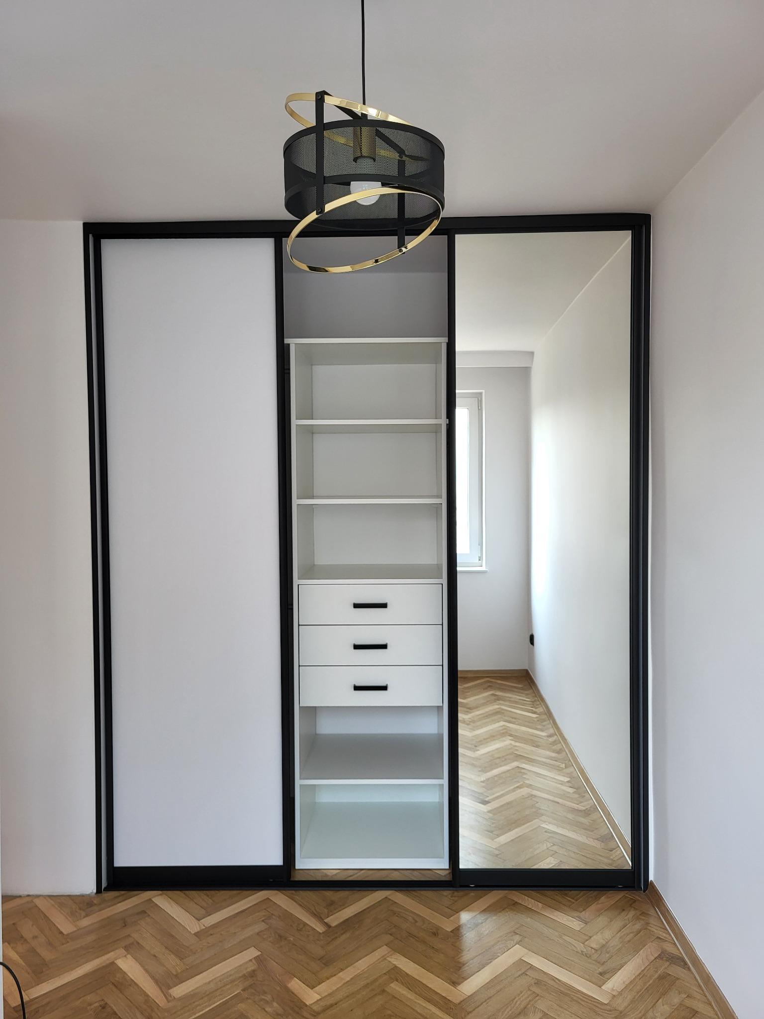 Nowoczesna szafa przesuwna z białymi frontami i czarną ramą, z lustrem na jednym skrzydle, widok na wnętrze pokoju z parkietem w jodełkę, oświetlona ozdobną lampą sufitową.