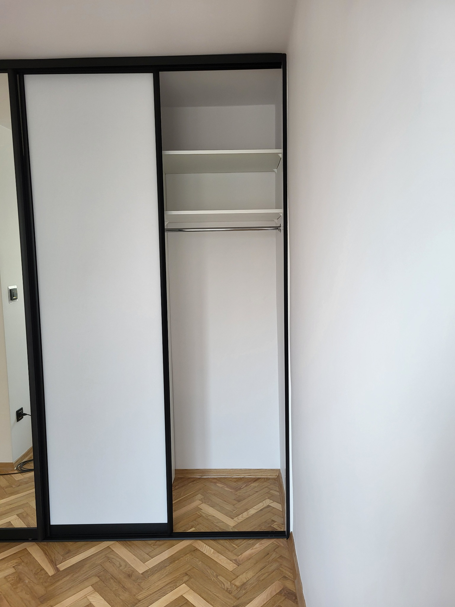 Biała szafa przesuwna z czarnymi obramowaniami, widoczne półki i drążek na ubrania, parkiet w jodełkę na podłodze.