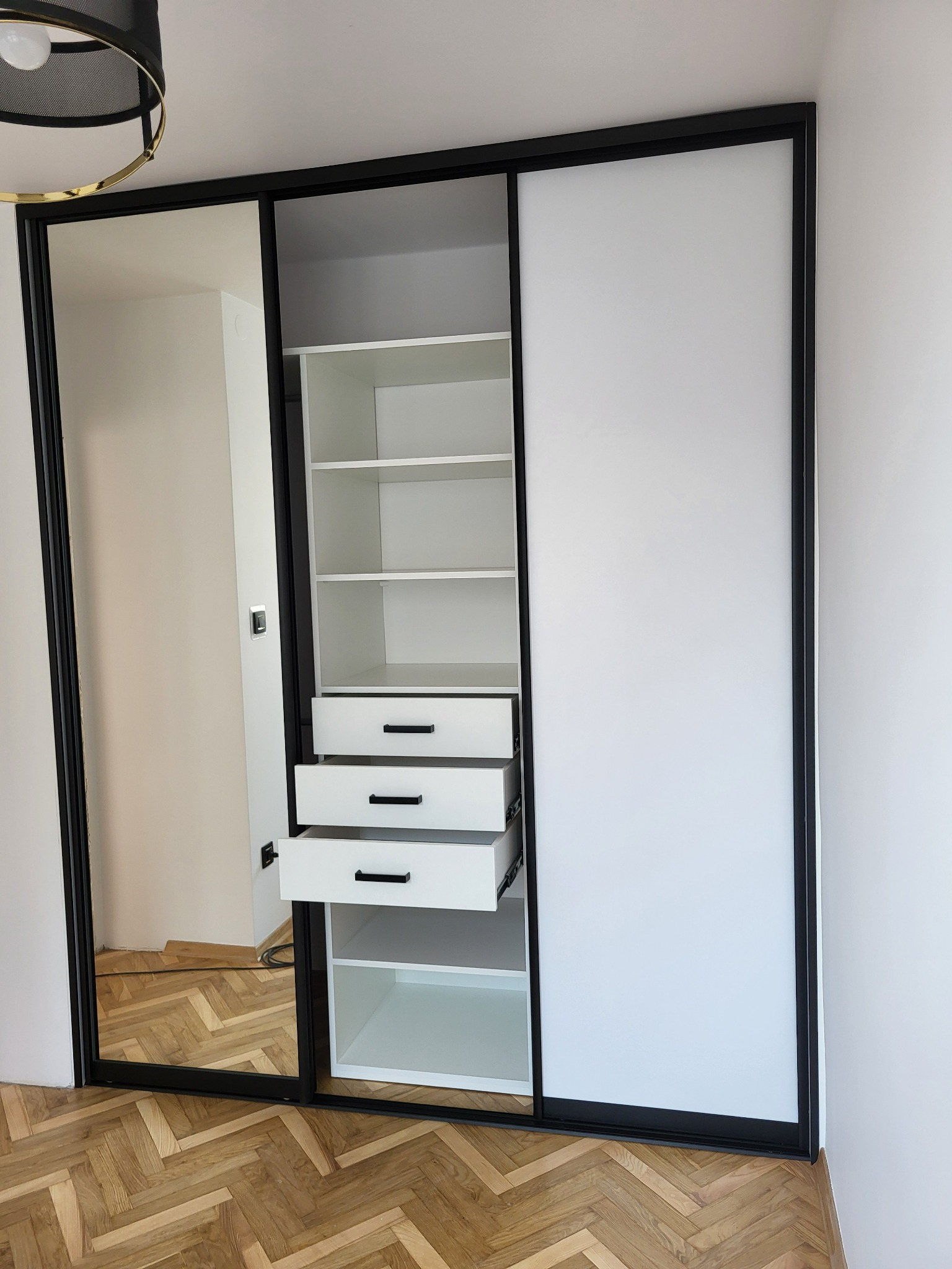 Wnętrze szafy przesuwnej z lustrem i białymi frontami, czarne obramowanie, widoczne półki i wysuwane szuflady z czarnymi uchwytami, parkiet w jodełkę na podłodze.