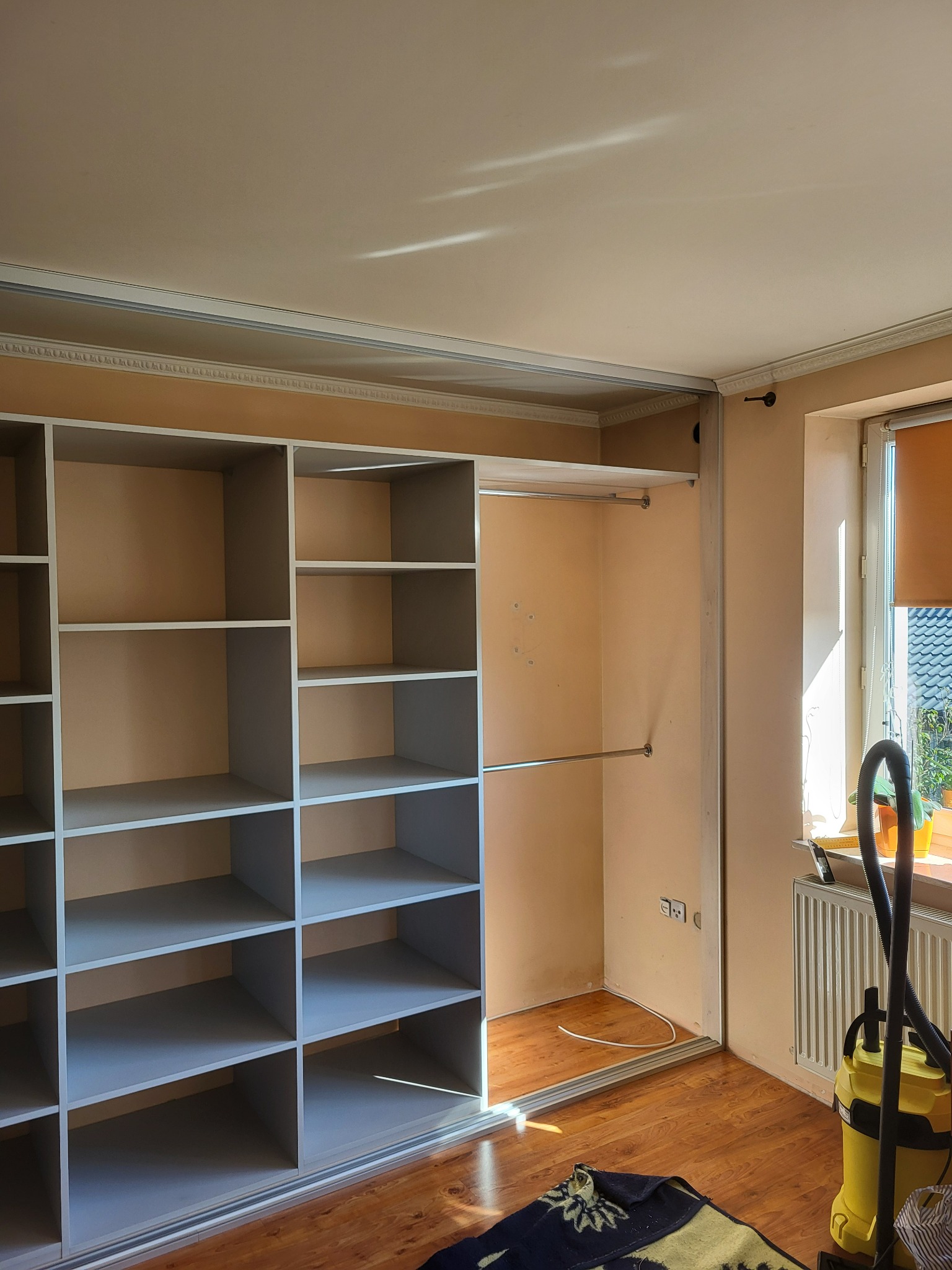 Wnętrze pokoju z częściowo zamontowaną, dużą szafą wnękową z przesuwnymi drzwiami i półkami, widoczne ślady po montażu i odkurzacz na tle okna.