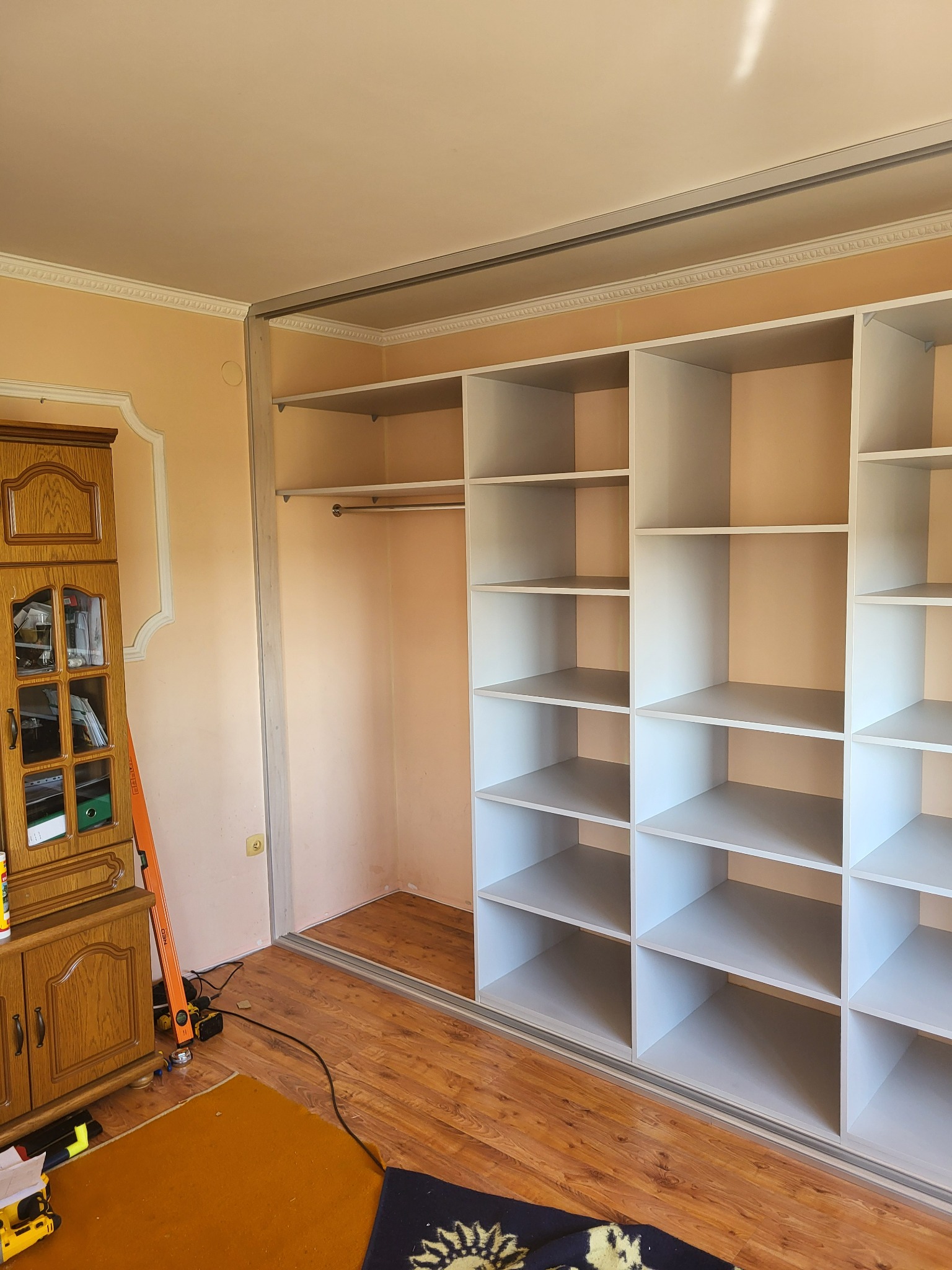 Wnętrze szafy wnękowej z białymi półkami, widoczny drążek na ubrania, obok stoi drewniany kredens i narzędzia pracy.