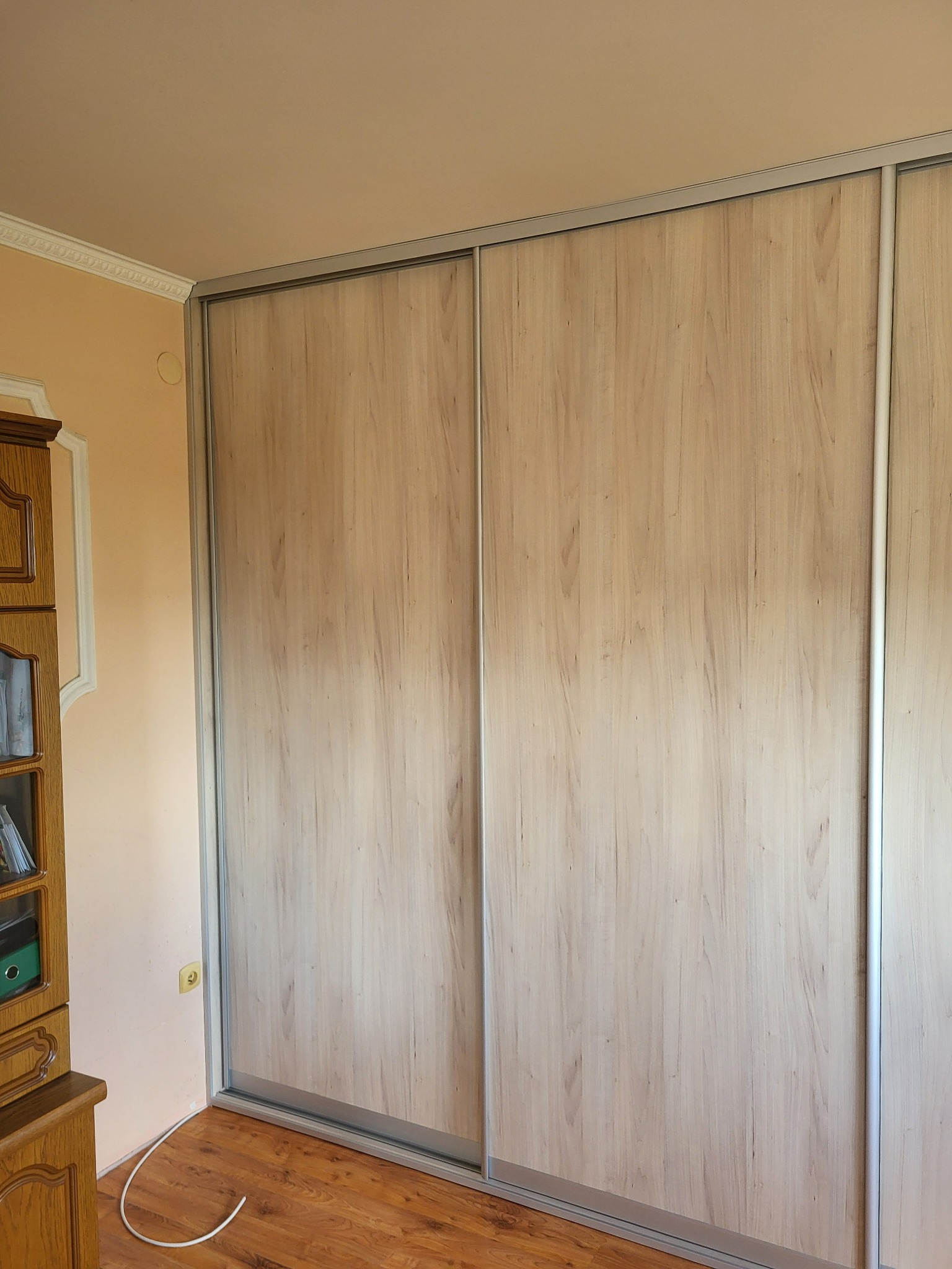 Zamknięta szafa z przesuwnymi drzwiami o jasnym, drewnopodobnym wykończeniu i srebrnych obramowaniach, widoczna w rogu pokoju z drewnianą podłogą i starą szafką.