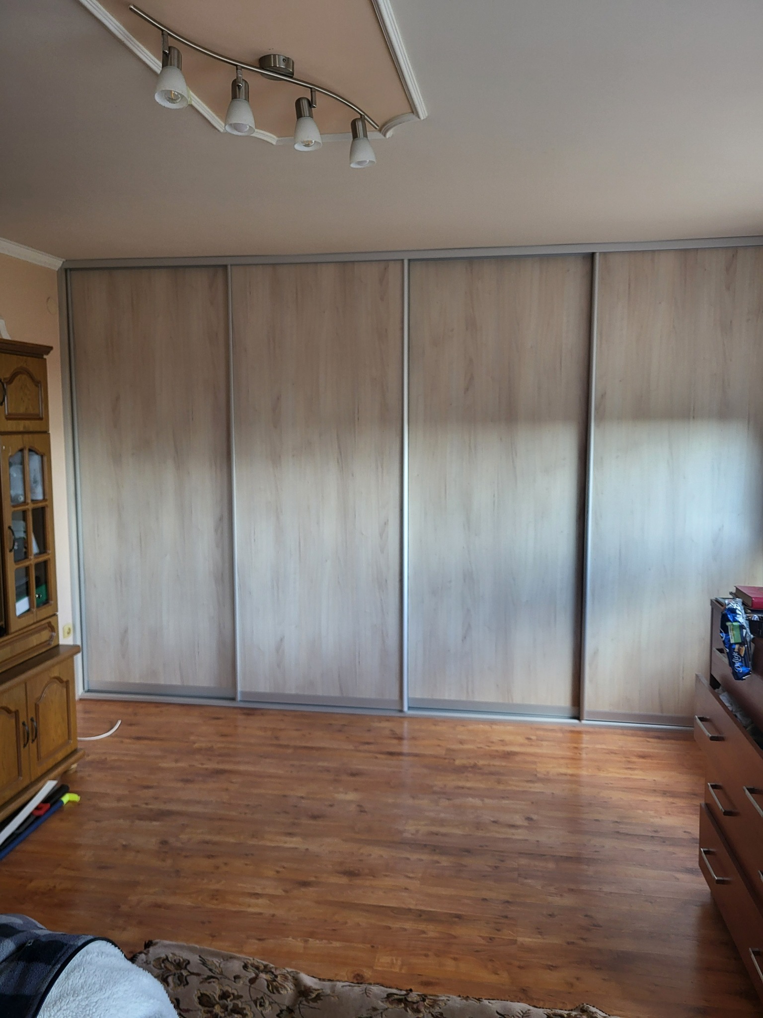 Duża szafa przesuwna z jasnego drewna z czterema drzwiami, zabudowana od ściany do ściany, widoczna na tle parkietu i fragmentów mebli.