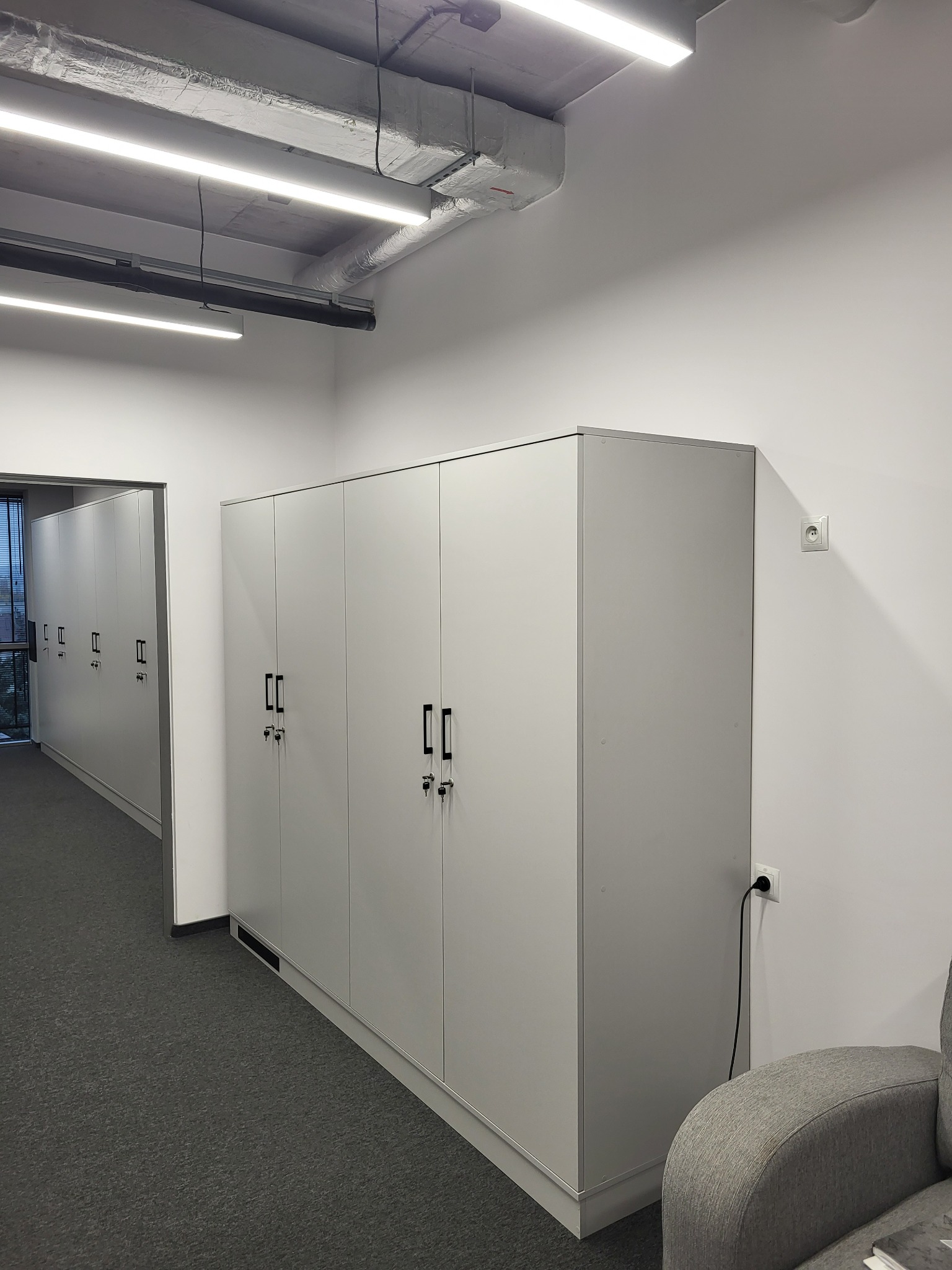 Białe szafy biurowe z czarnymi uchwytami i zamkami na klucz, ustawione w rzędzie wzdłuż ściany w Woli Rzędzińskiej, widoczne instalacje pod sufitem.