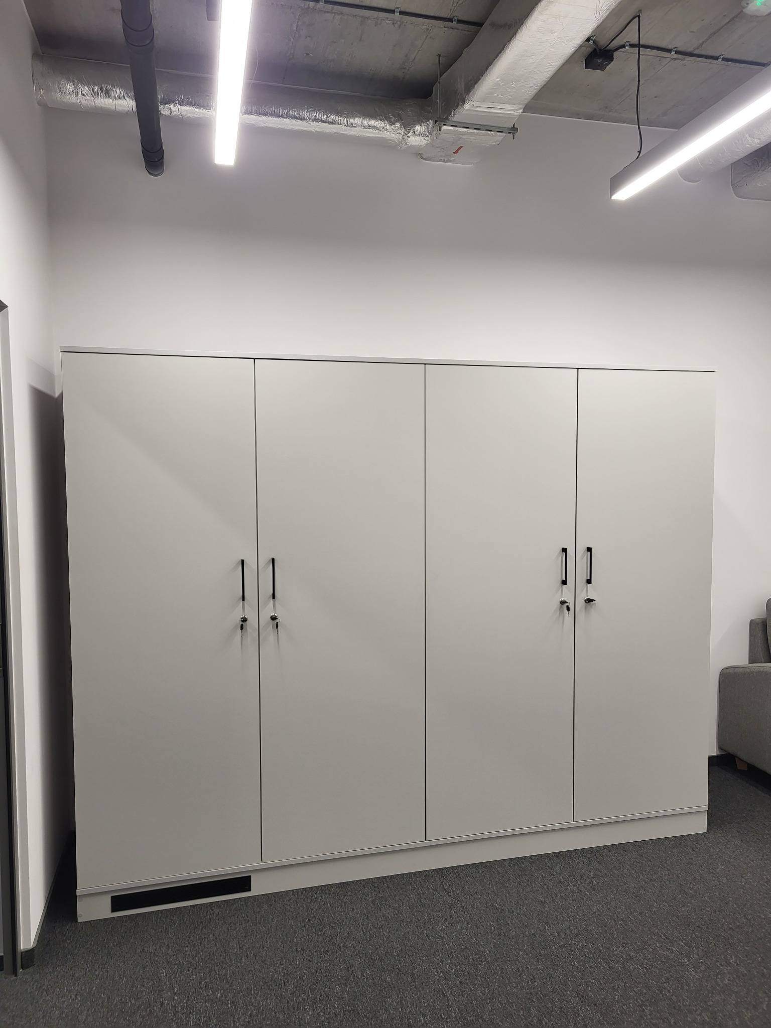 Biała, minimalistyczna szafa z czterema drzwiami i czarnymi uchwytami, z kluczami w zamkach, na tle szarej wykładziny w nowoczesnym biurze z widocznymi elementami wentylacji pod sufitem.