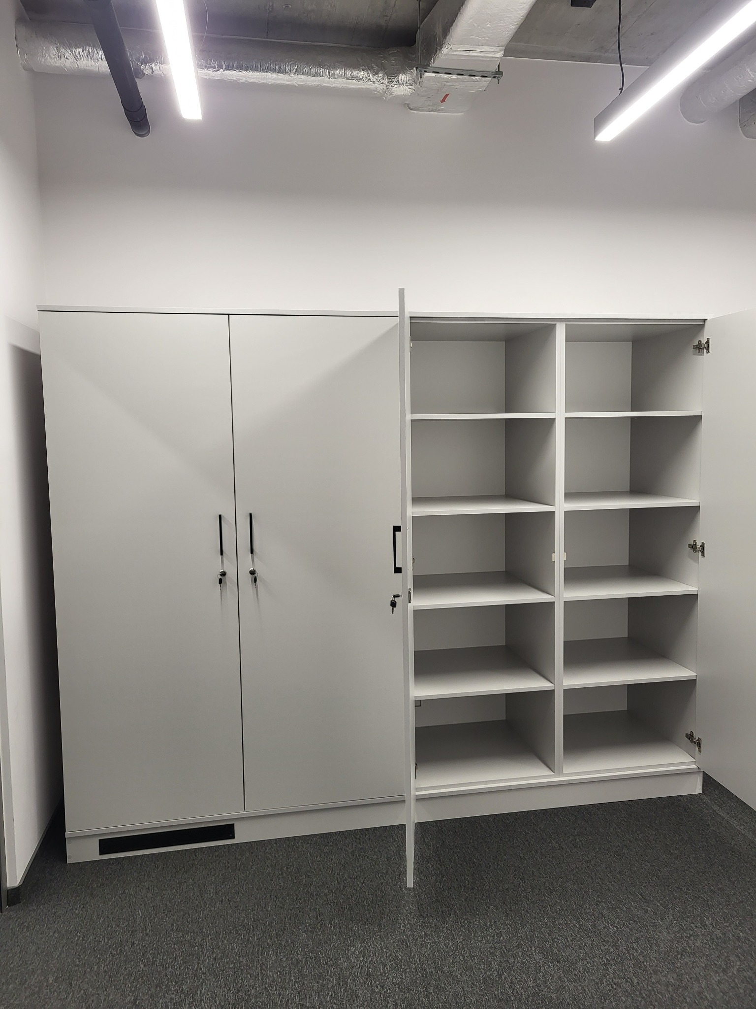 Biała szafa biurowa z dwiema parami drzwi zamykanymi na klucz i otwartą częścią regałową z półkami, widoczna instalacja wentylacyjna pod sufitem.