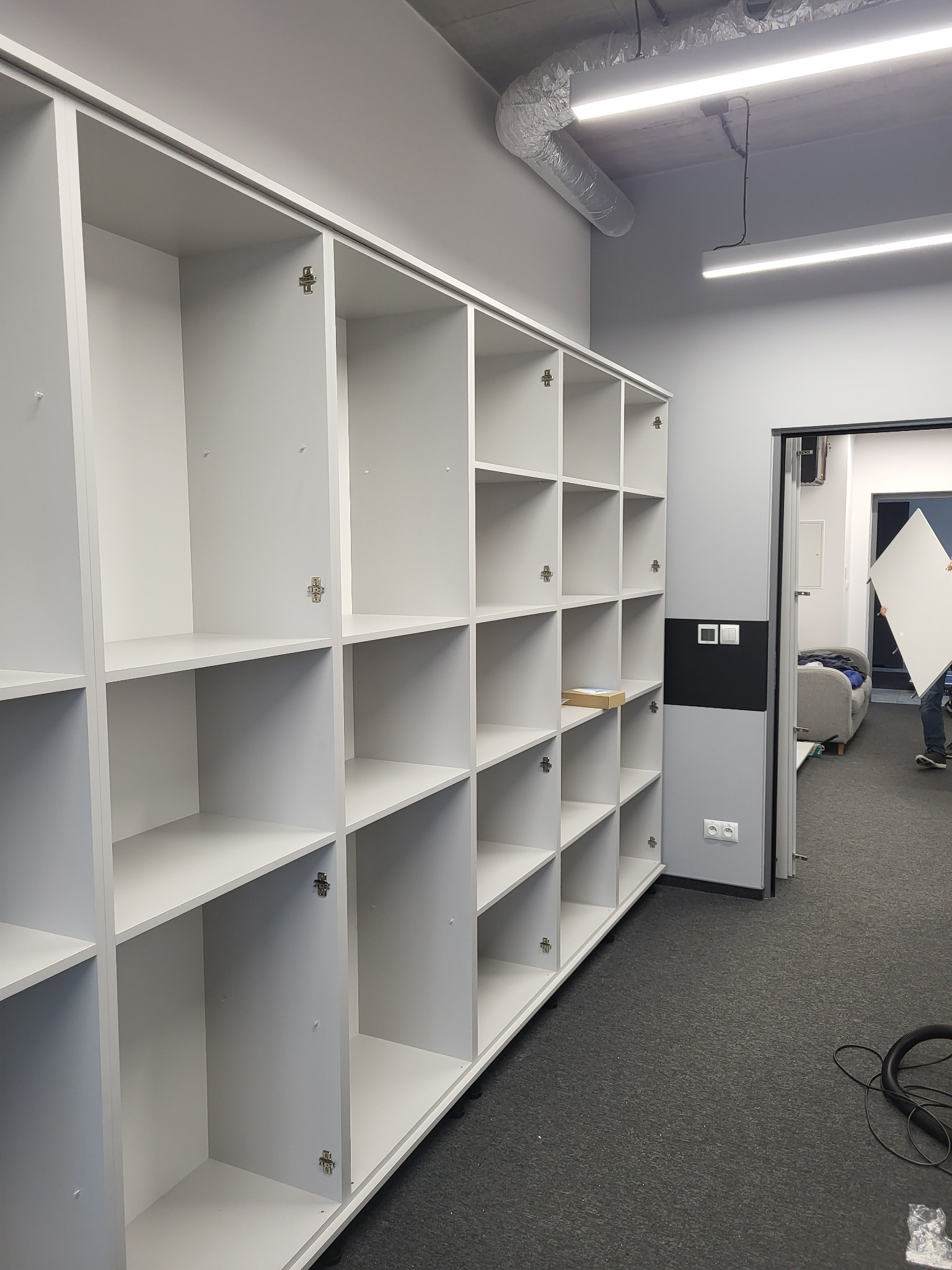 Biała, modułowa konstrukcja regałowa z otwartymi półkami, widoczne zawiasy, w tle osoba przenosząca białą płytę, szara wykładzina podłogowa i fragment odkurzacza.