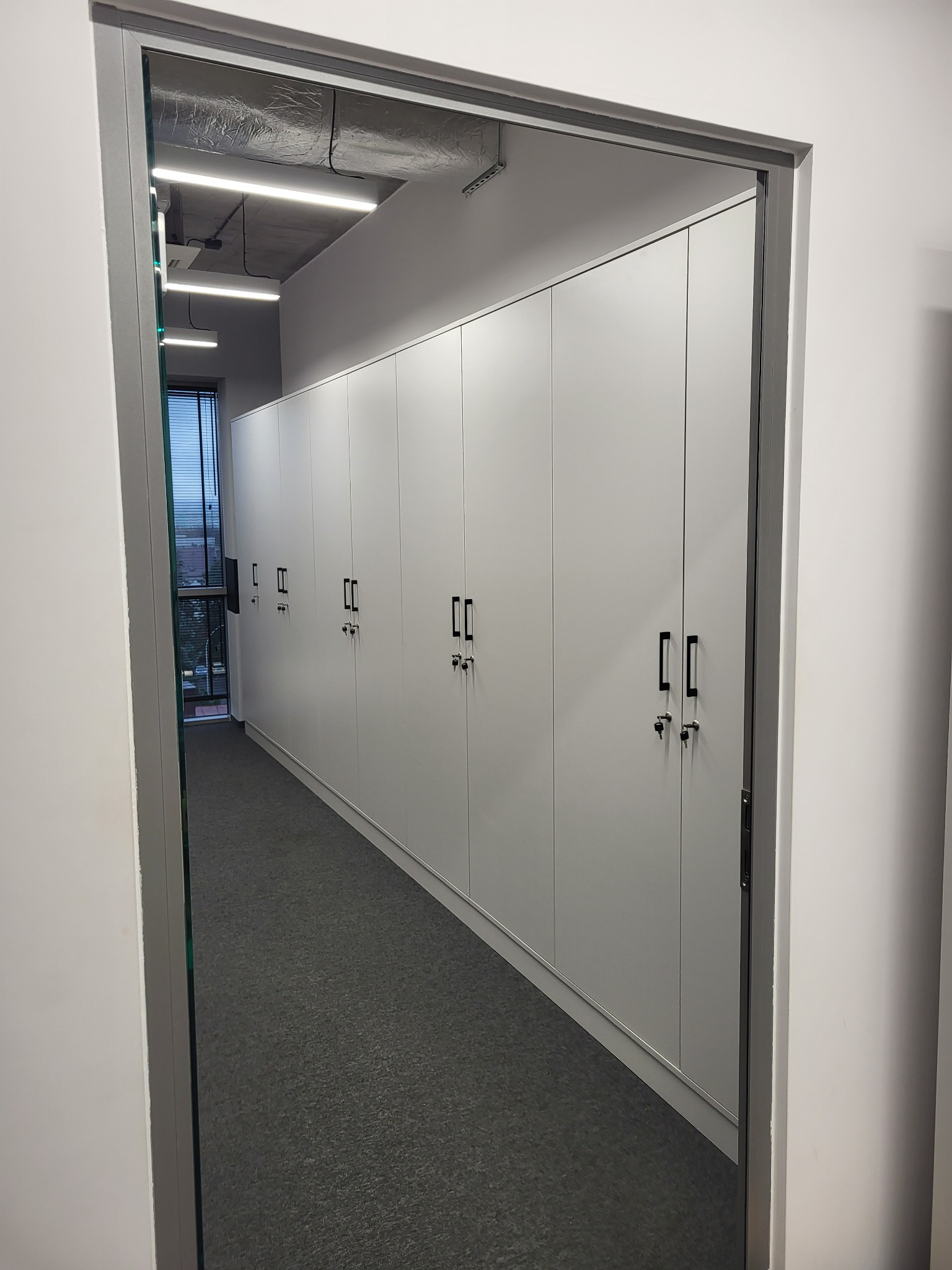 Długi rząd białych szaf z czarnymi uchwytami i zamkami, widoczny zza framugi drzwiowej w biurze z szarą wykładziną.