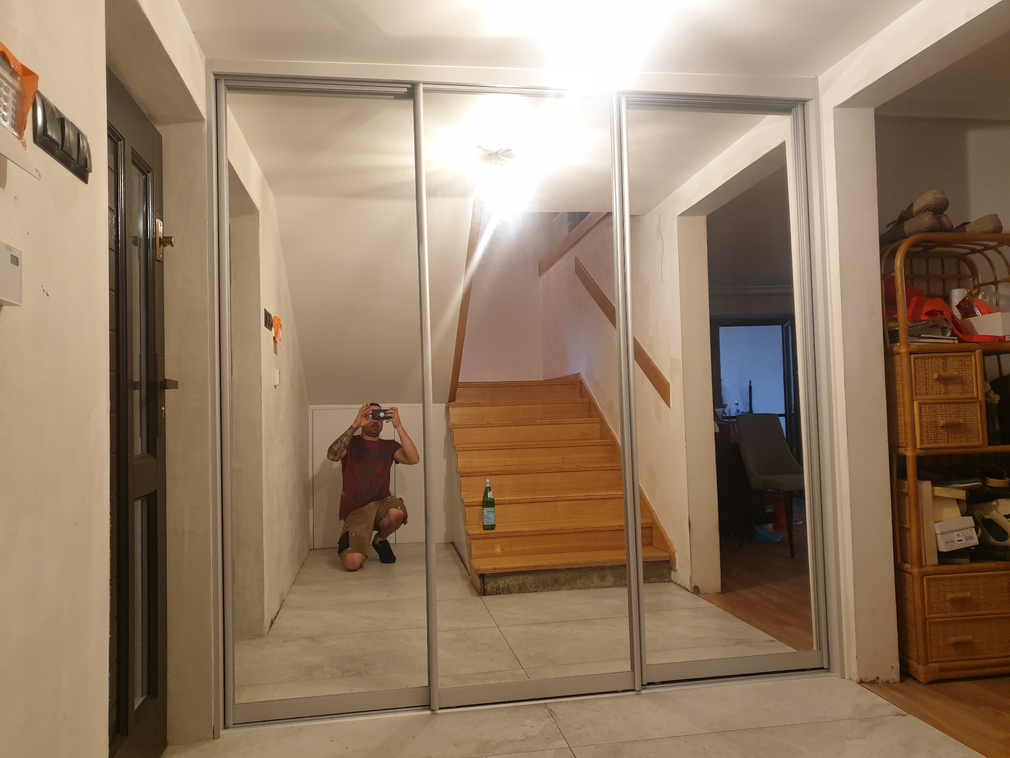 Wnętrze z trzydrzwiową szafą przesuwną z lustrzanymi frontami, odbijającą klatkę schodową i osobę robiącą zdjęcie telefonem.