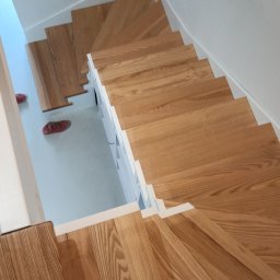 KRZYSZTOF JAGIELNIK - Drewniane schody z białymi podstopnicami widziane z góry, z parą czerwonych butów i stopą w skarpetce z motywem Batmana na dolnym planie.