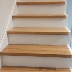 KRZYSZTOF JAGIELNIK - Jasne drewniane schody z białymi podstopnicami, widok z dołu, naturalne oświetlenie eksponuje usłojenie drewna.