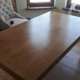 Rozkładany stół z drewna o ciepłym odcieniu, z widocznym usłojeniem, ustawiony w jasnym wnętrzu z drewnianą podłogą i krzesłem w stylu vintage w tle.