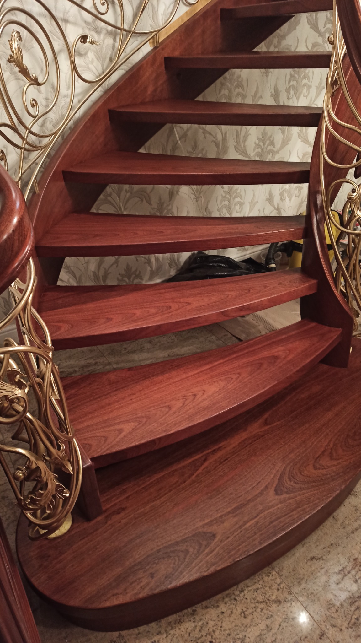 Klatka schodowa z drewna o ciepłym, czerwono-brązowym odcieniu, z ozdobnymi, metalowymi balustradami w kolorze złota, widok od dołu, na tle wzorzystej tapety.