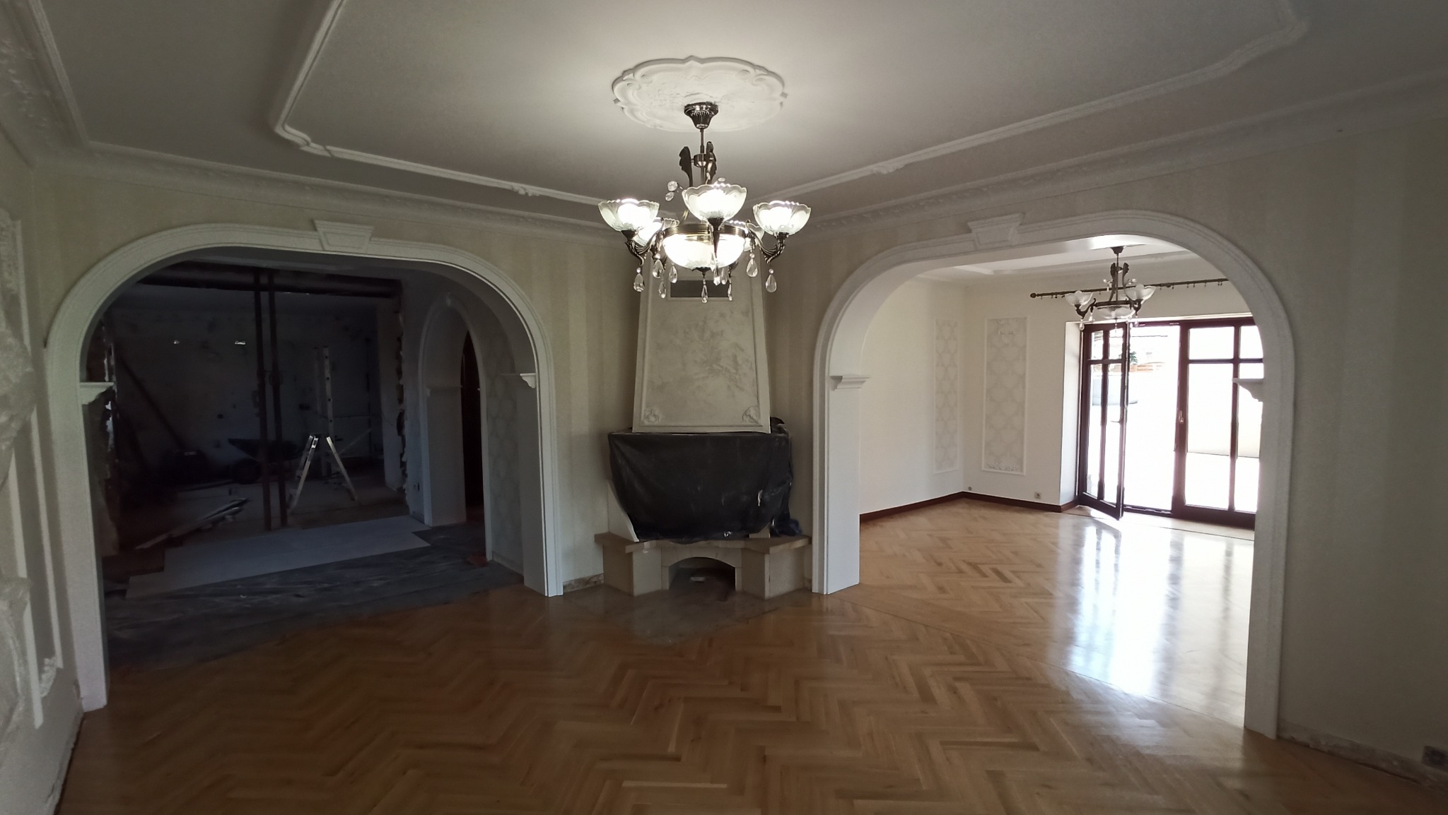 Salon z parkietem ułożonym w jodełkę, widoczne przejścia w formie łuków, żyrandol z kryształkami i kominek zasłonięty czarną folią, w tle widoczne drzwi balkonowe i pomieszczenie w trakcie remontu.