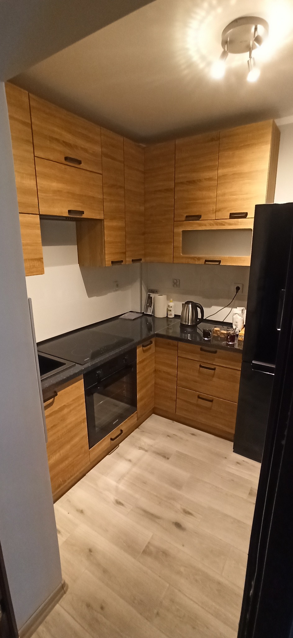 Kuchnia z drewnianymi szafkami i czarnym blatem, widoczny fragment lodówki i podłoga z paneli imitujących drewno.