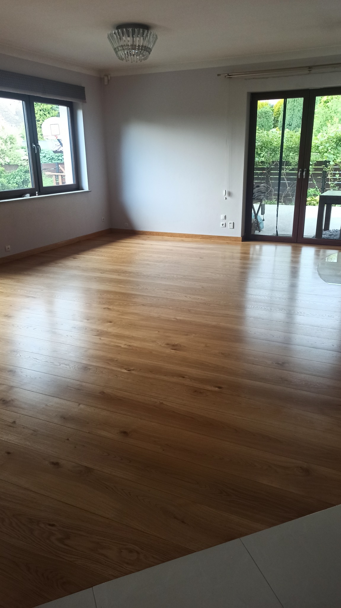 Jasny, przestronny pokój z nową, lśniącą podłogą z drewnianych desek ułożonych w poprzek, widok na ogród przez duże okno i drzwi balkonowe.