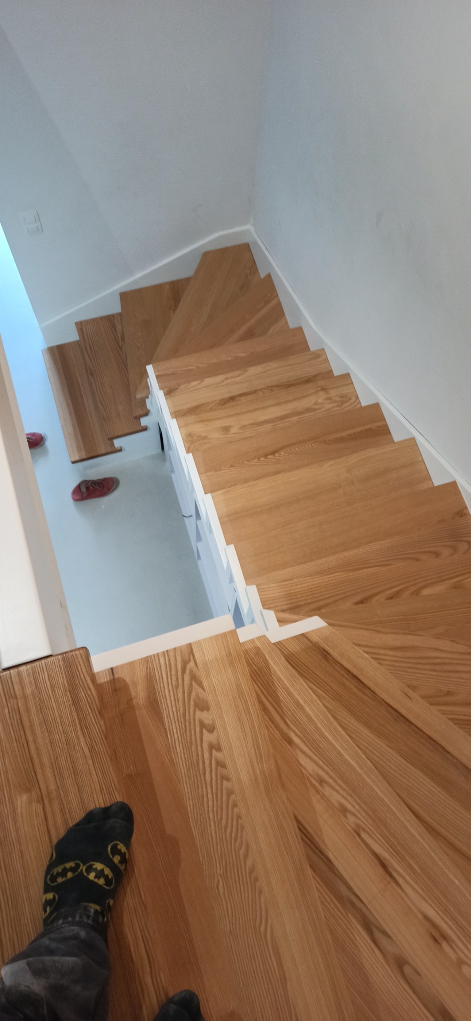 Drewniane schody z białymi podstopnicami widziane z góry, z parą czerwonych butów i stopą w skarpetce z motywem Batmana na dolnym planie.
