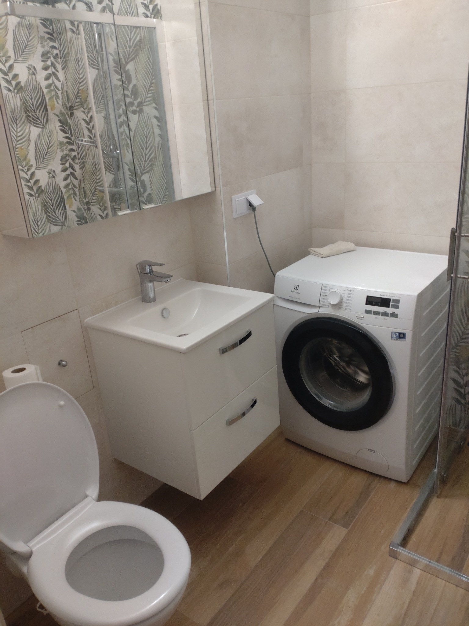 Jasna łazienka z umywalką na szafce, toaletą i pralką Electrolux, wykończona beżowymi płytkami i drewnopodobną podłogą, widok na kabinę prysznicową.