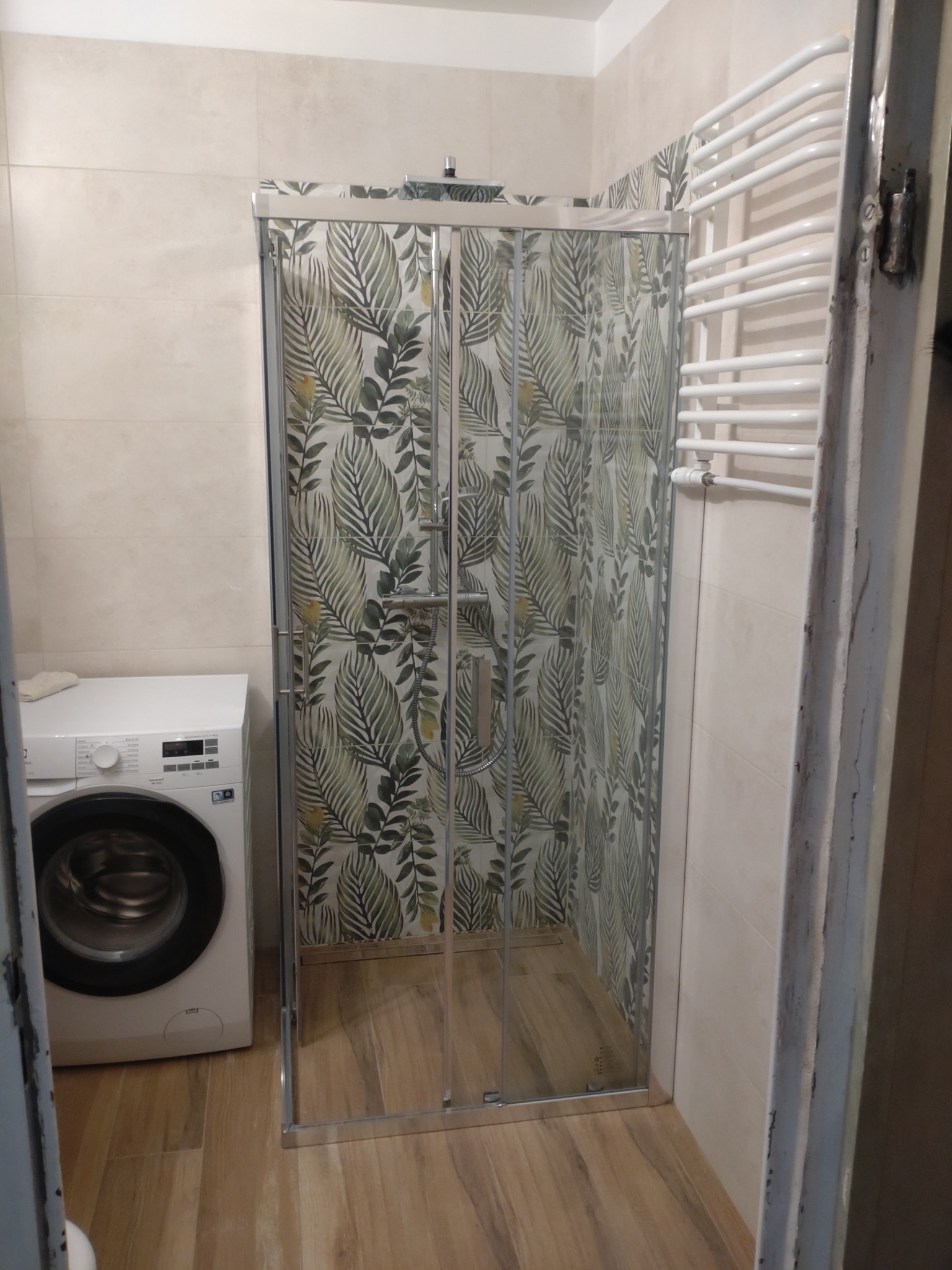 Nowoczesna kabina prysznicowa z wzorem liści na ścianie, obok pralka i grzejnik drabinkowy w jasnej łazience z drewnopodobną podłogą.