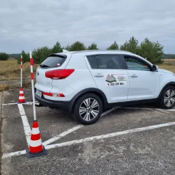 Biały samochód osobowy marki Kia na placu manewrowym z pachołkami i słupkami, widoczny logotyp ośrodka szkolenia kierowców El-Goral na drzwiach.