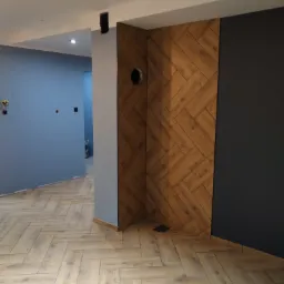 Świeżo wykończone wnętrze z podłogą ułożoną w jodełkę, drewnopodobna okładzina ściany w tym samym wzorze oraz fragment pomalowanej na ciemny kolor ściany, widoczne przygotowane otwory instalacyjne.