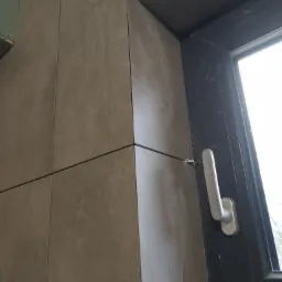 Szare płytki ceramiczne ułożone wokół ciemnego okna z białą klamką, widoczne fugi i ślady po pracach wykończeniowych.