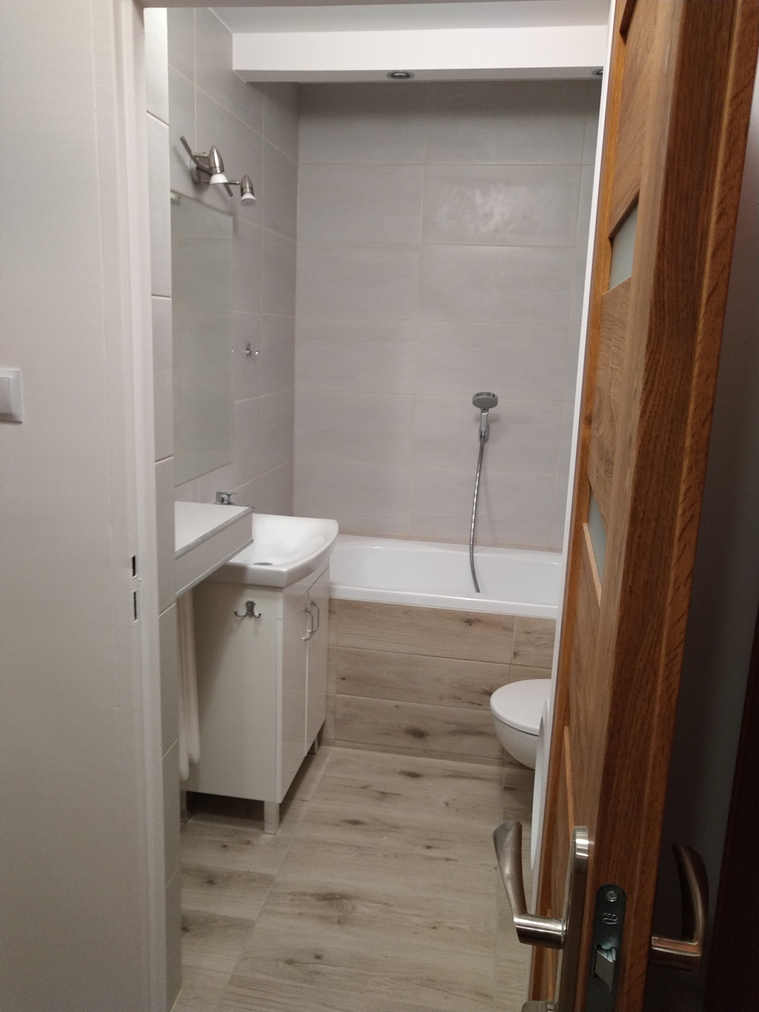 Wykończona łazienka z jasnymi płytkami, drewnopodobną zabudową wanny i podłogą, białą szafką pod umywalką oraz drewnianymi drzwiami z przeszkleniami.