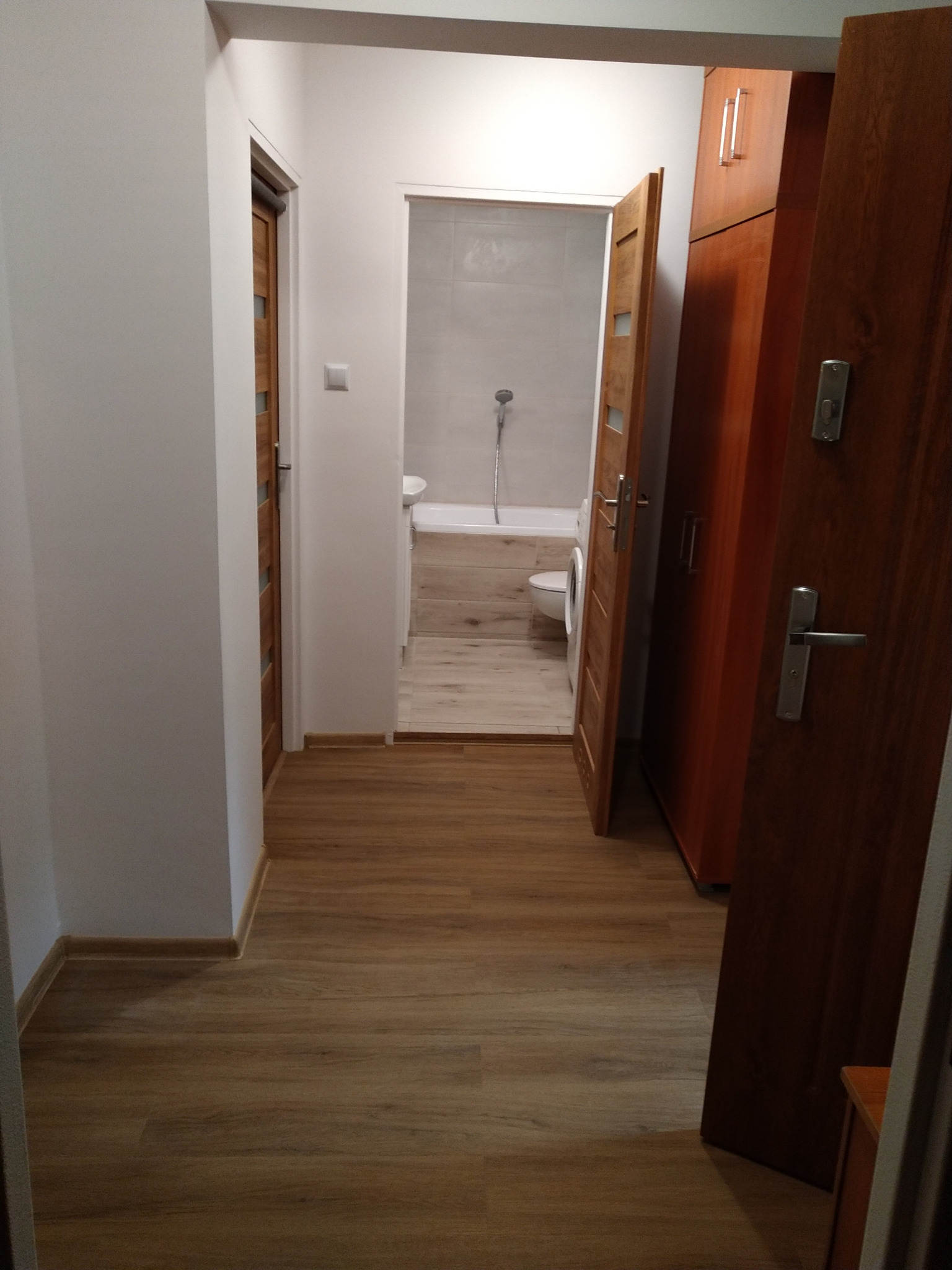 Widok z korytarza na wykończoną łazienkę z drewnopodobną podłogą, białą wanną i toaletą, z częściowo otwartymi drewnianymi drzwiami i szafą w tle.