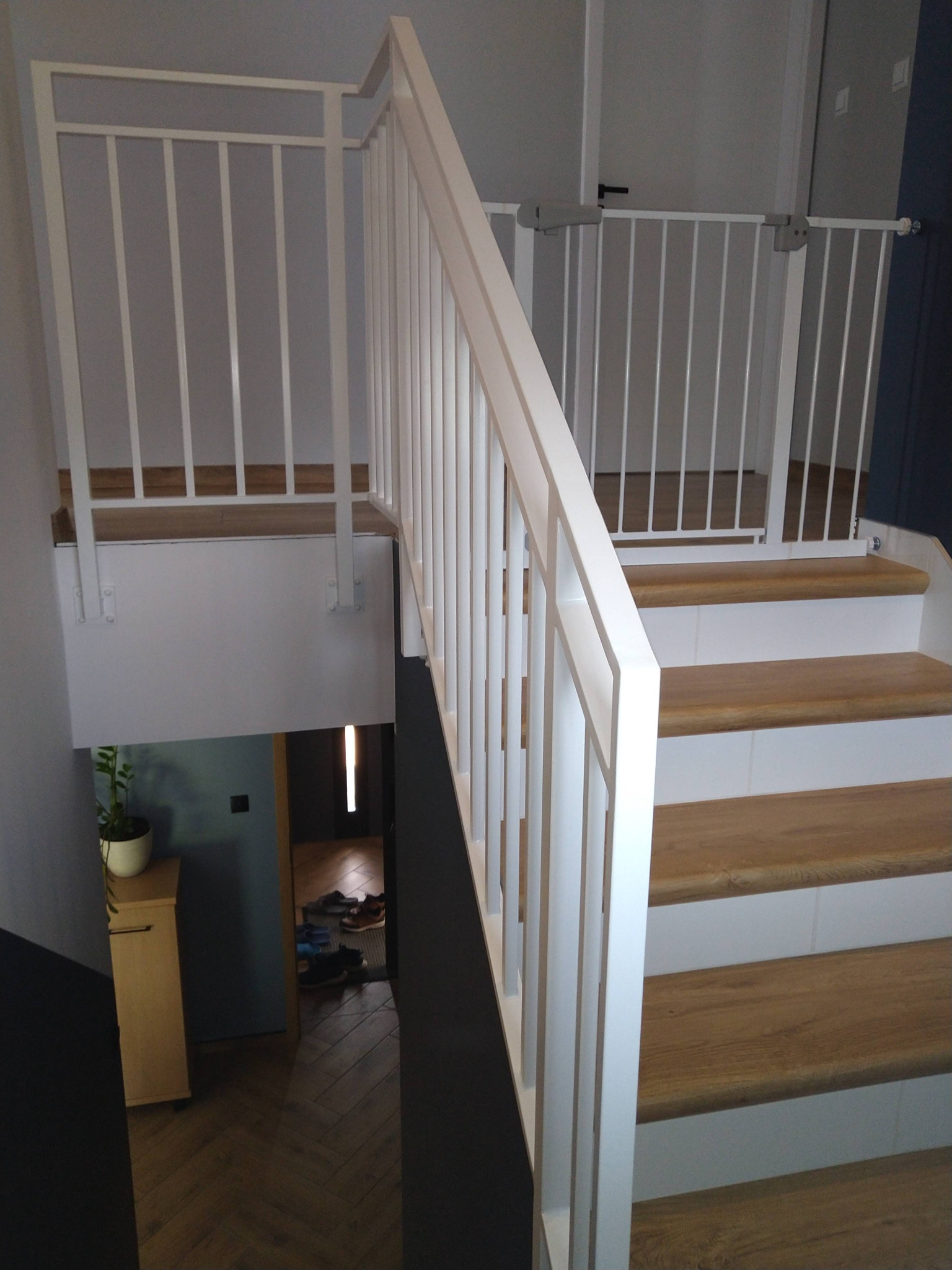 Białe schody z drewnianymi stopniami i białą balustradą, zabezpieczone białą bramką dla dziecka na górze, widok na dół z szafką i rośliną w doniczce.