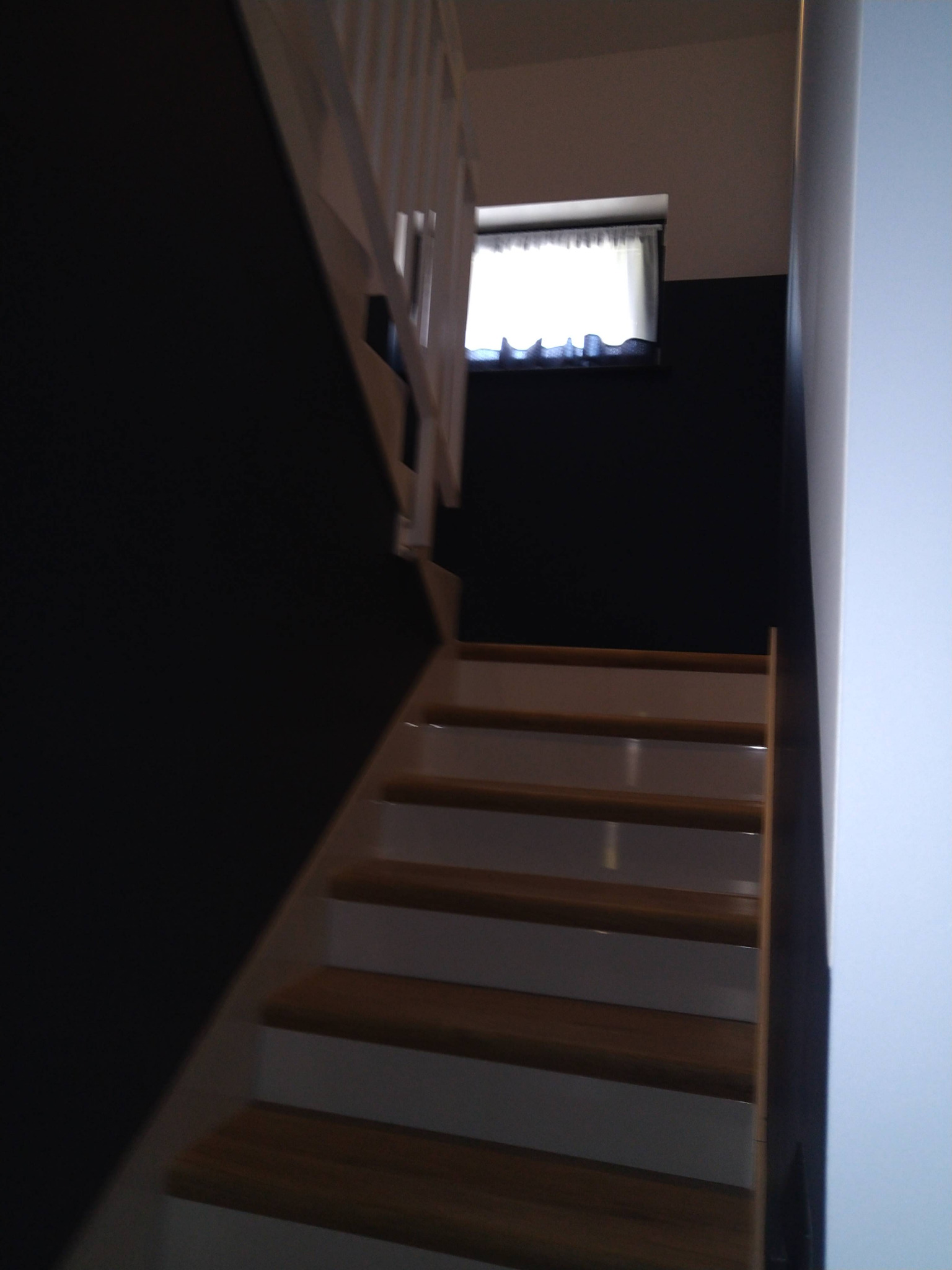 Schody drewniane z białymi elementami na tle ciemnej ściany, widoczne okno z zasłoną.