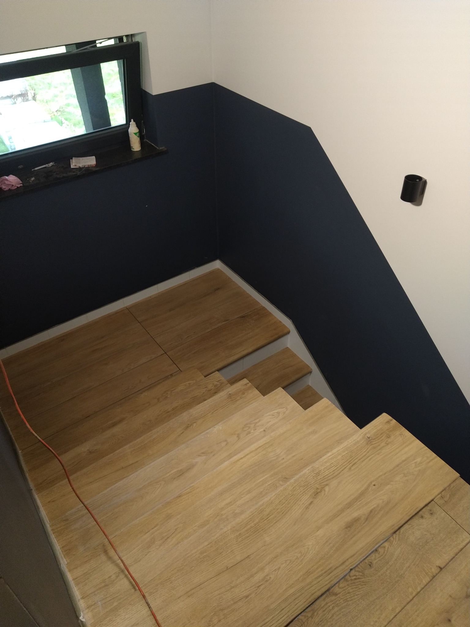 Montaż drewnianych paneli na schodach w Kędzierzynie-Koźlu, widoczne niedomalowane ściany w kolorze granatowym i białym, oraz fragment okna.