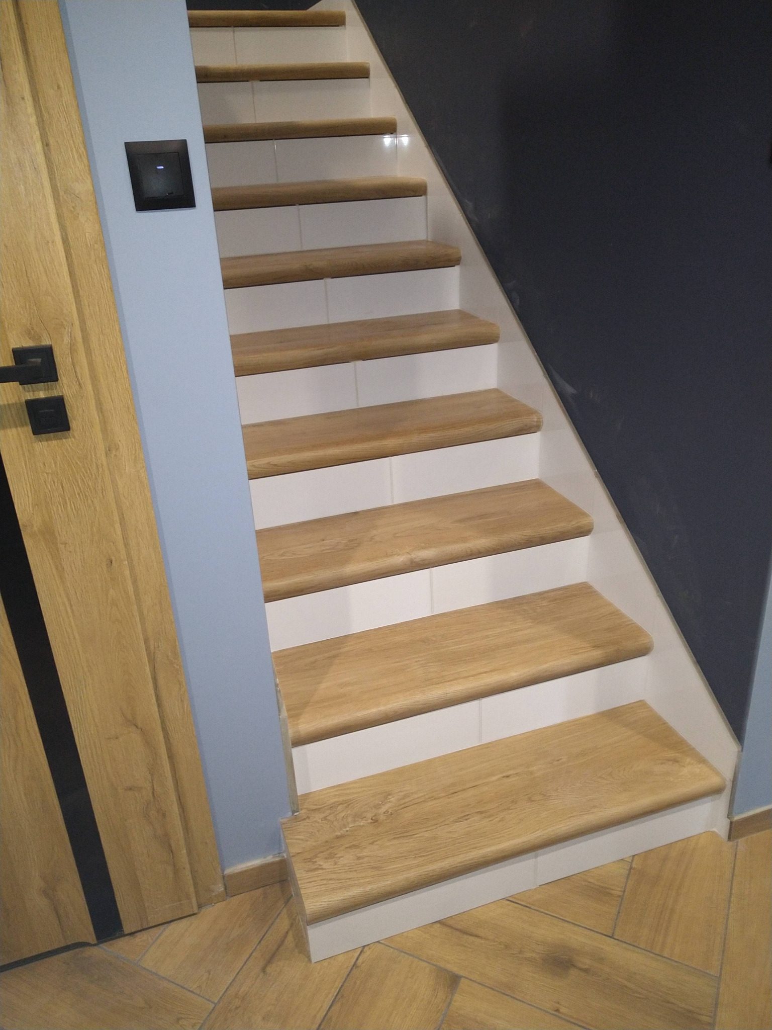 Drewniane schody z białymi podstopnicami, przylegające do szarej ściany, z widoczną drewnianą podłogą i drzwiami z czarnymi akcentami.