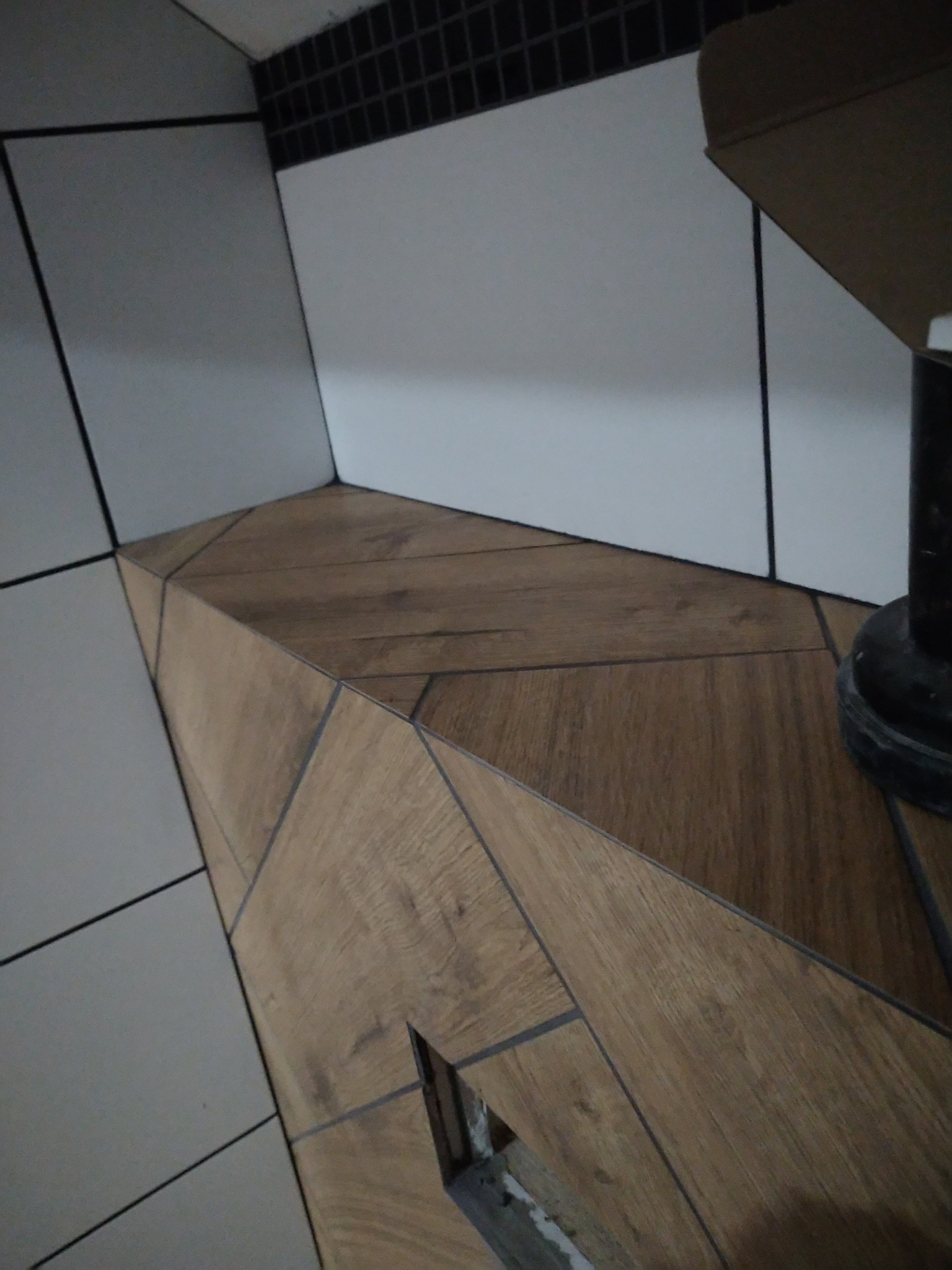 Nietypowe ułożenie płytek imitujących drewno na obudowie w łazience, widoczne białe płytki na ścianach z czarną fugą, niedokończone wykończenie w dolnej części obudowy.