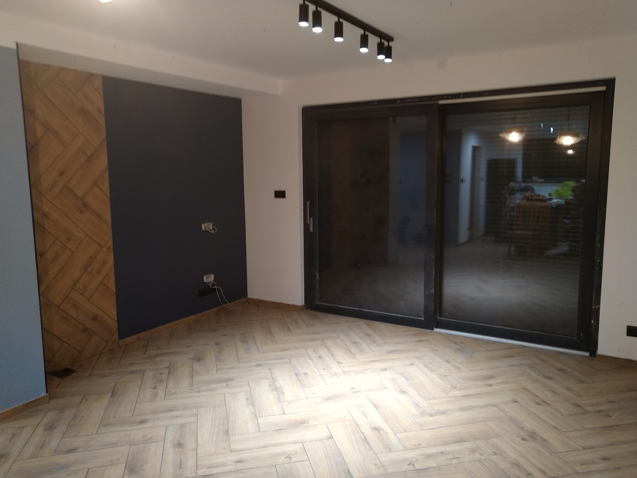 Wykończone wnętrze pokoju z parkietem ułożonym w jodełkę, ciemną ścianą z drewnianą wstawką i przesuwnymi drzwiami z czarną ramą, oświetlonym listwą z punktowymi lampami.
