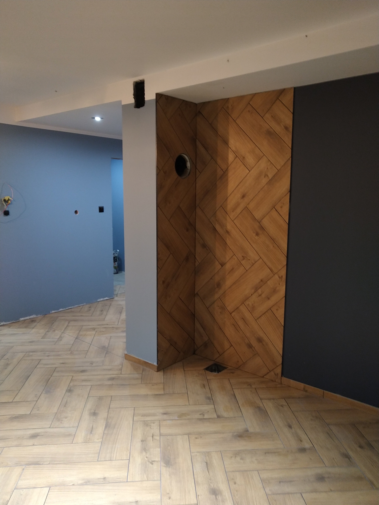 Świeżo wykończone wnętrze z podłogą ułożoną w jodełkę, drewnopodobna okładzina ściany w tym samym wzorze oraz fragment pomalowanej na ciemny kolor ściany, widoczne przygotowane otwory instalacyjne.