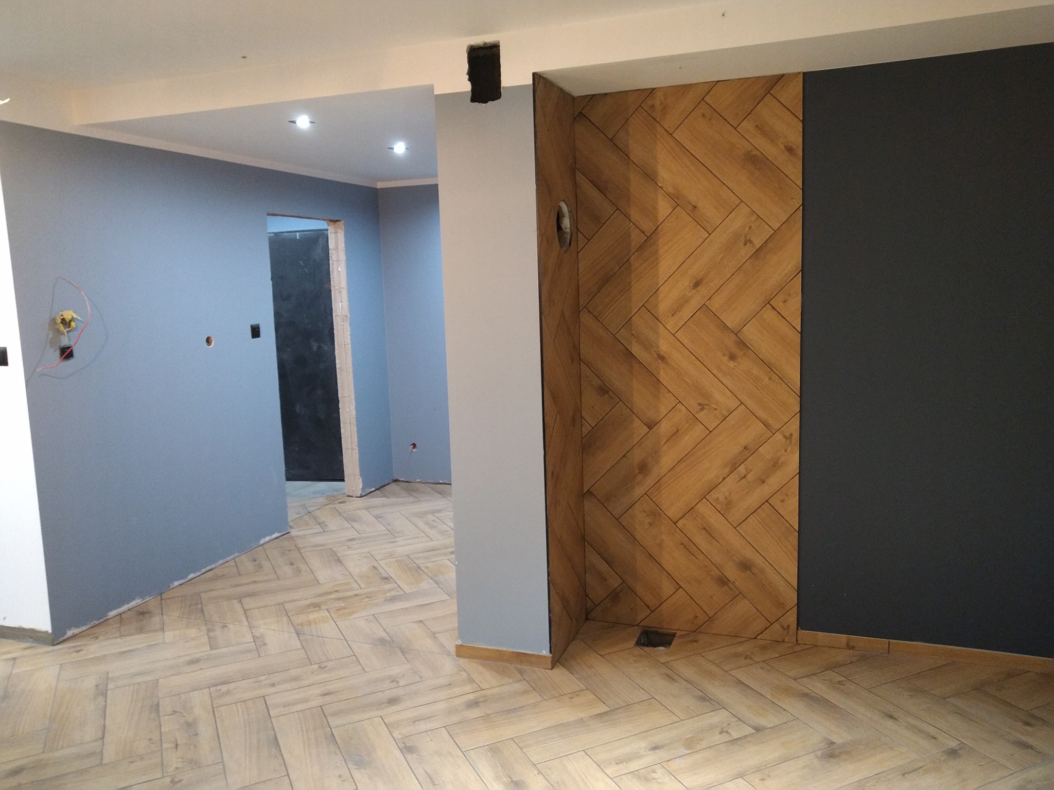 Wnętrze w trakcie prac wykończeniowych: szara ściana, drewnopodobna podłoga ułożona w jodełkę, ściana z drewnianych paneli w jodełkę oraz fragment ściany pomalowanej na ciemny kolor.