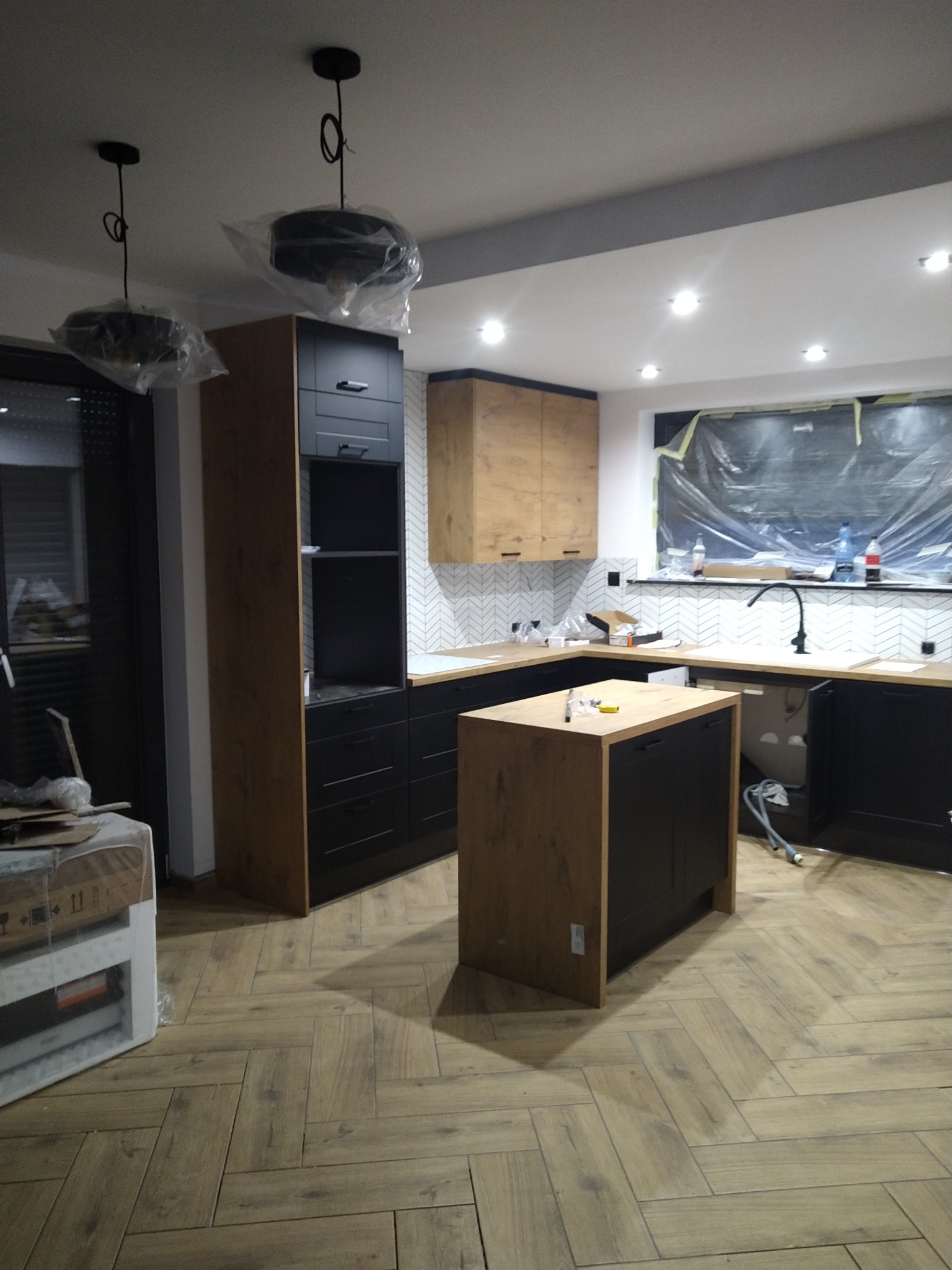 Montaż zabudowy kuchennej z wyspą w trakcie realizacji, widoczne zabezpieczenia okna folią, podłoga ułożona w jodełkę.