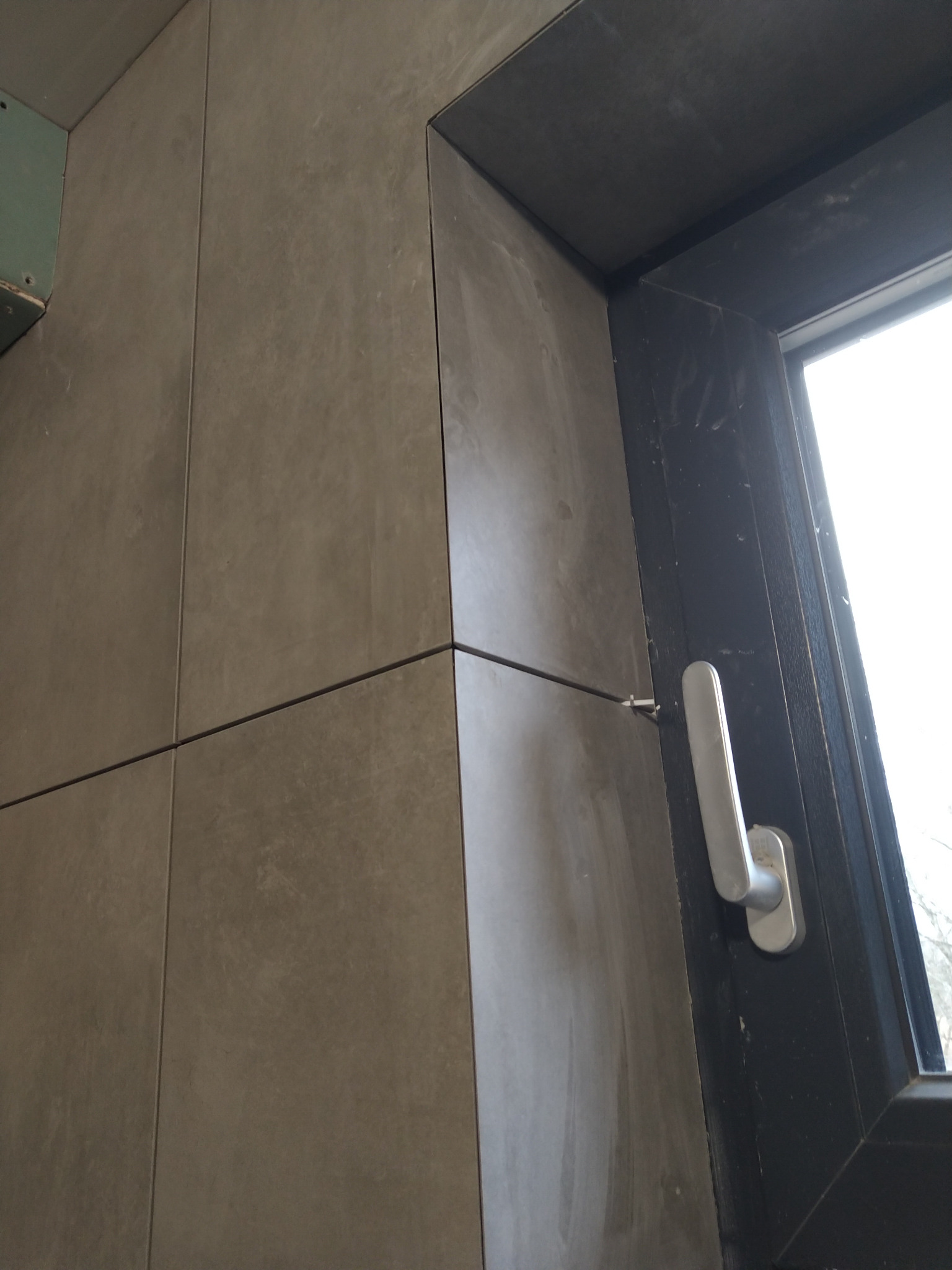 Szare płytki ceramiczne ułożone wokół ciemnego okna z białą klamką, widoczne fugi i ślady po pracach wykończeniowych.