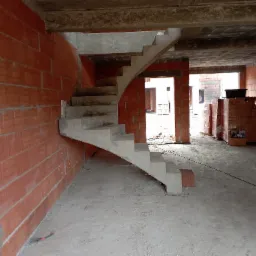 Surowe wnętrze w trakcie budowy: ściany z czerwonej cegły, betonowe schody łukowe prowadzące na wyższą kondygnację, widoczne materiały budowlane i otwory drzwiowe w tle.